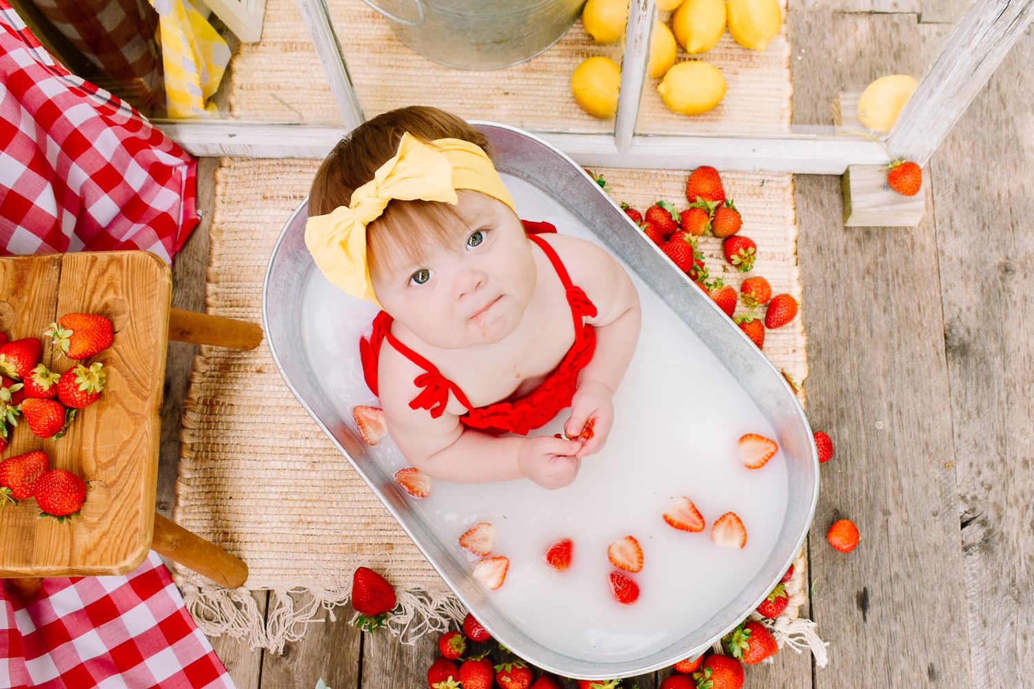 Baby girl with yellow bow sitting in milk bath tub surrounded by fresh strawberries and lemons.