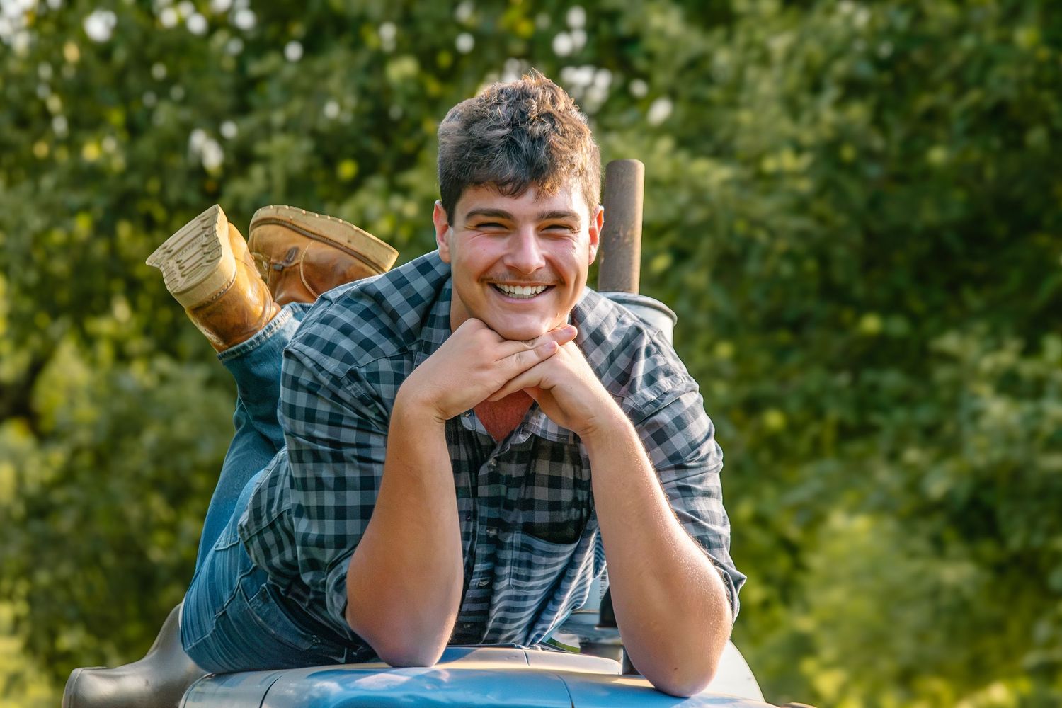 Smiling young man in plaid shirt leaning on car roof outdoors with cowboy boots visible, surrounded by green trees.