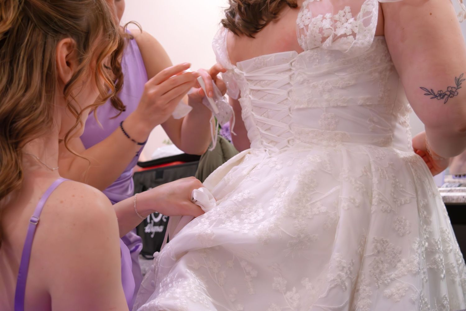 Bridesmaids in lavender dresses assisting bride with lacing up her elegant white wedding dress.