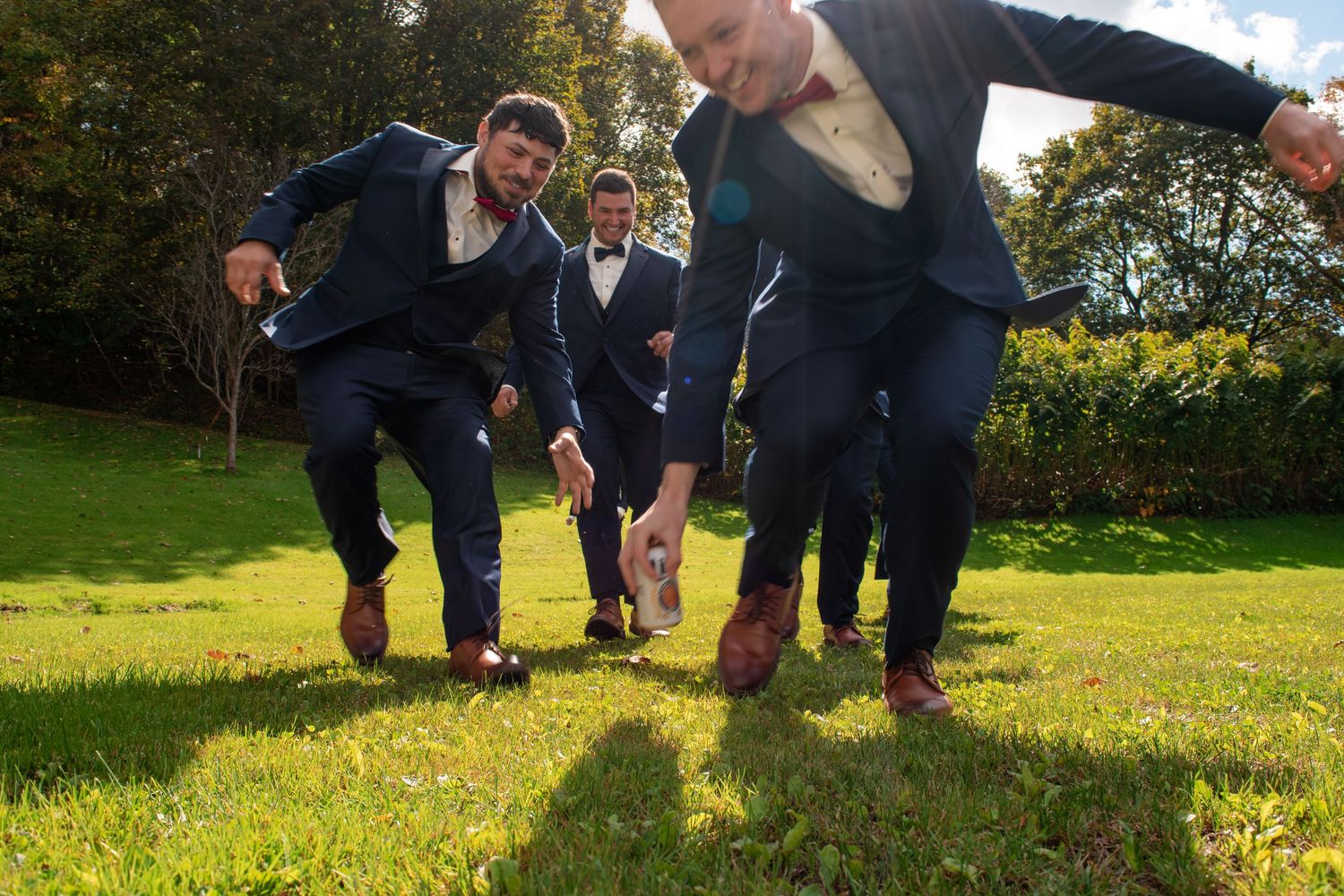 Groomsmen in navy suits playfully running on green grass outdoors at a wedding celebration.