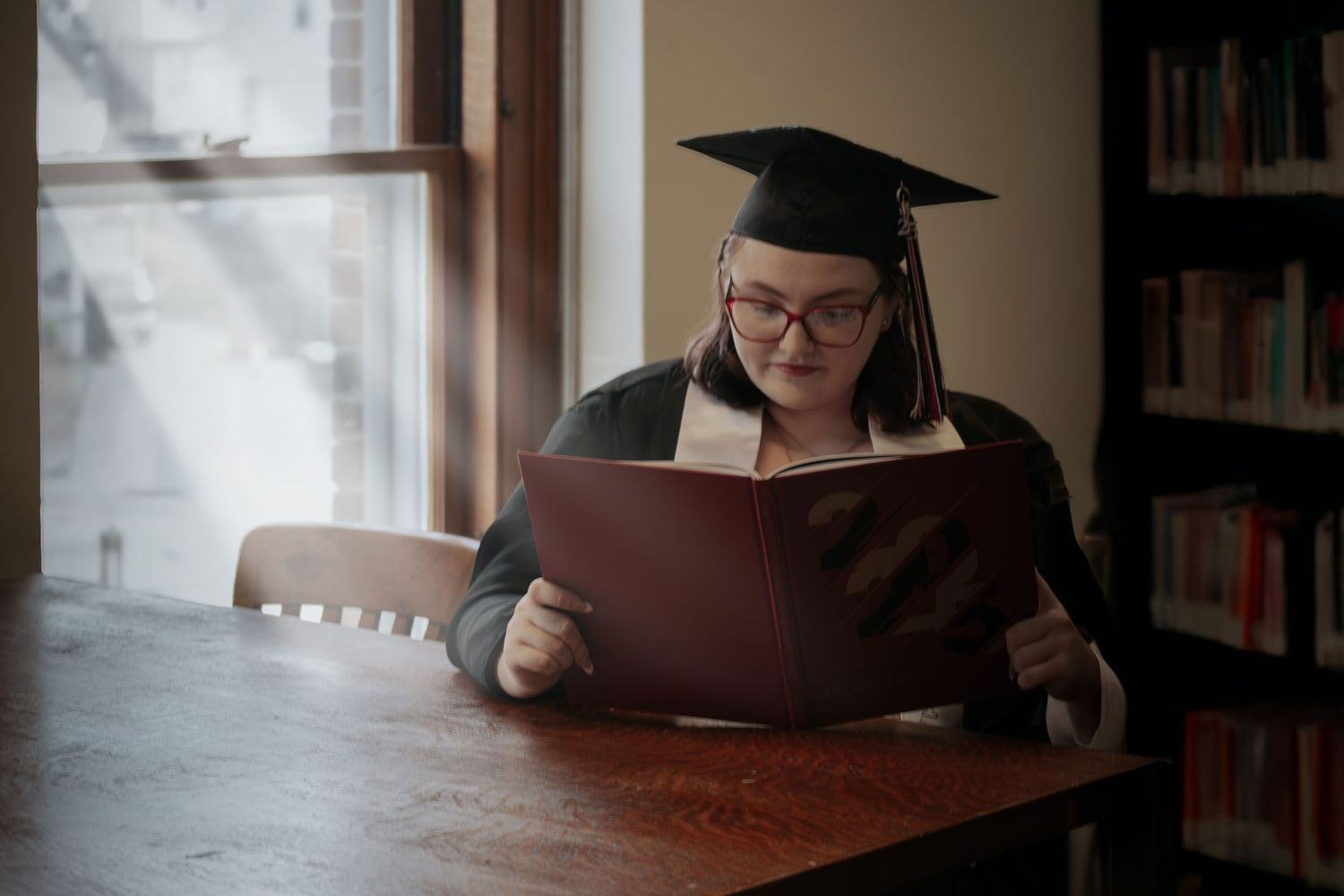 Graduate in cap and gown reading book at library table near window.