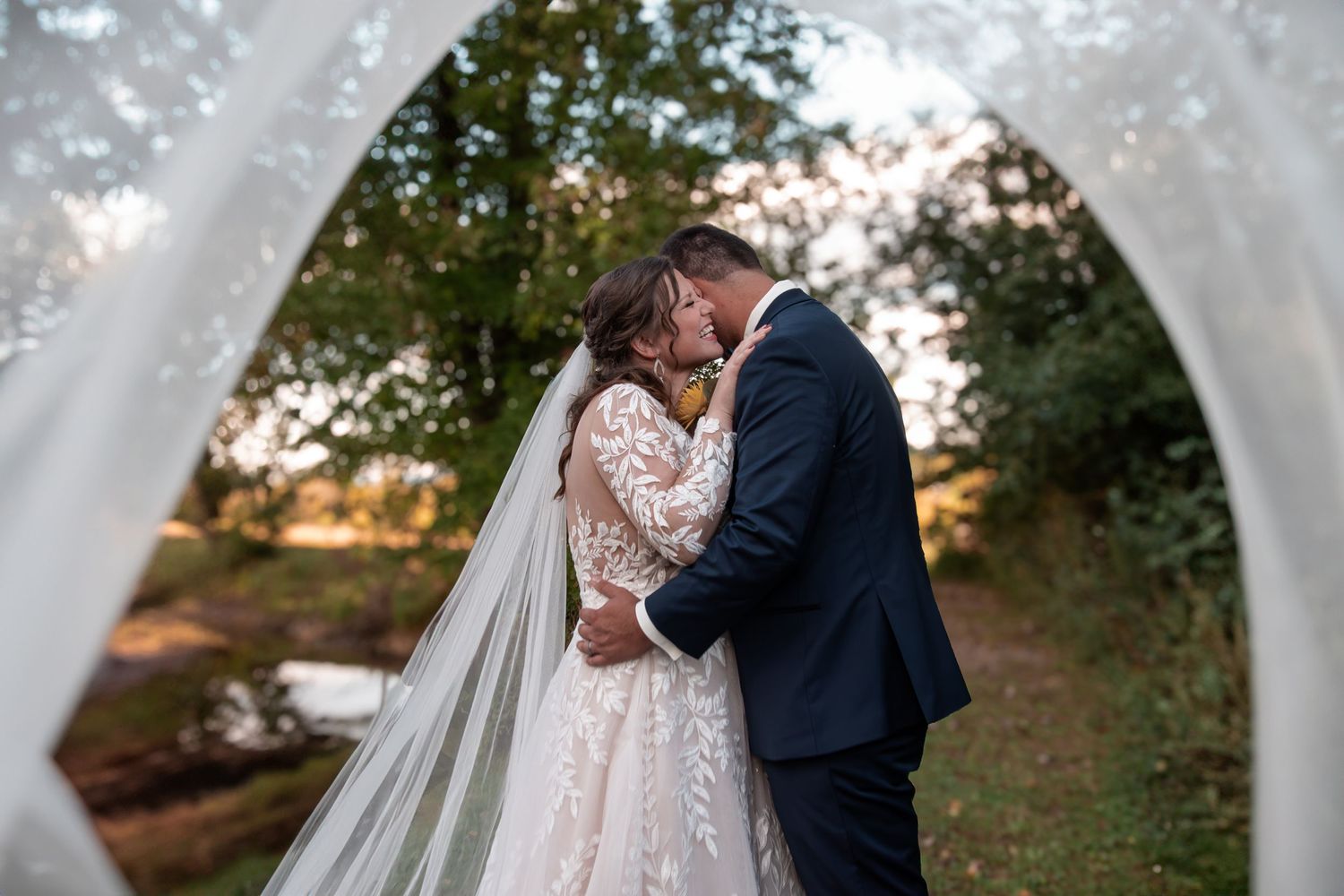 Bride and groom kiss framed by a floral arch draped with white tulle in a romantic outdoor autumn setting.