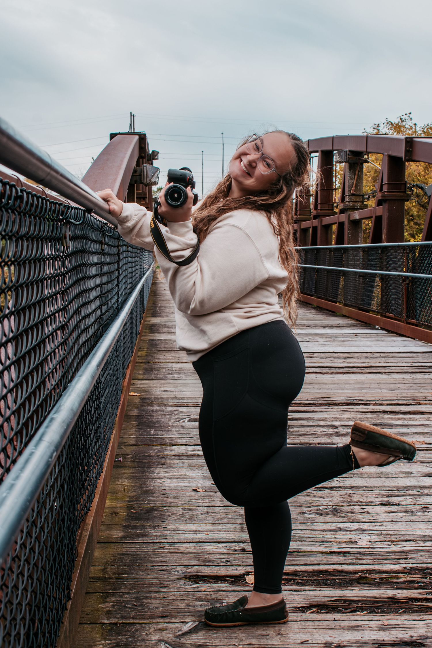 Happy woman posing with a camera on one leg on a rustic wooden bridge in autumn.