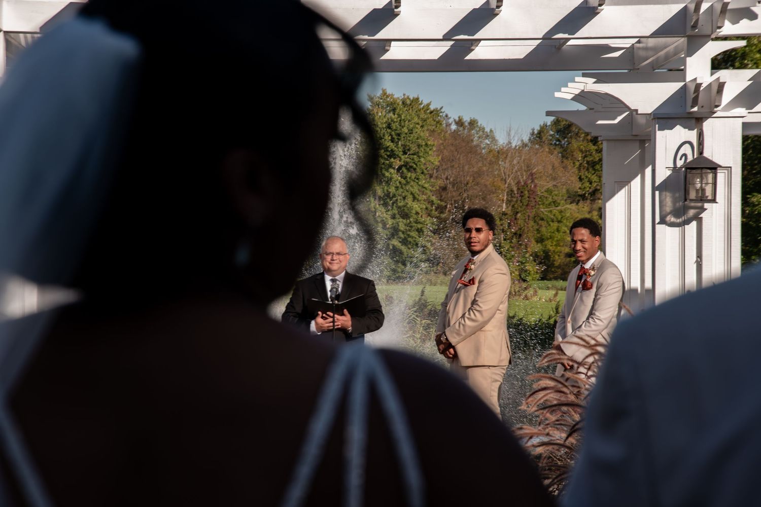 Groom and groomsmen in tan suits stand at outdoor wedding altar as bride approaches in white gown.