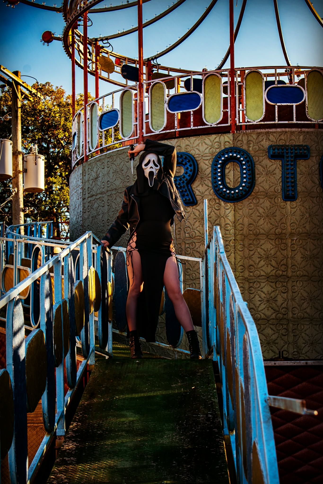 Person in Ghostface mask and black costume on fairground walkway near colorful Rotor ride.