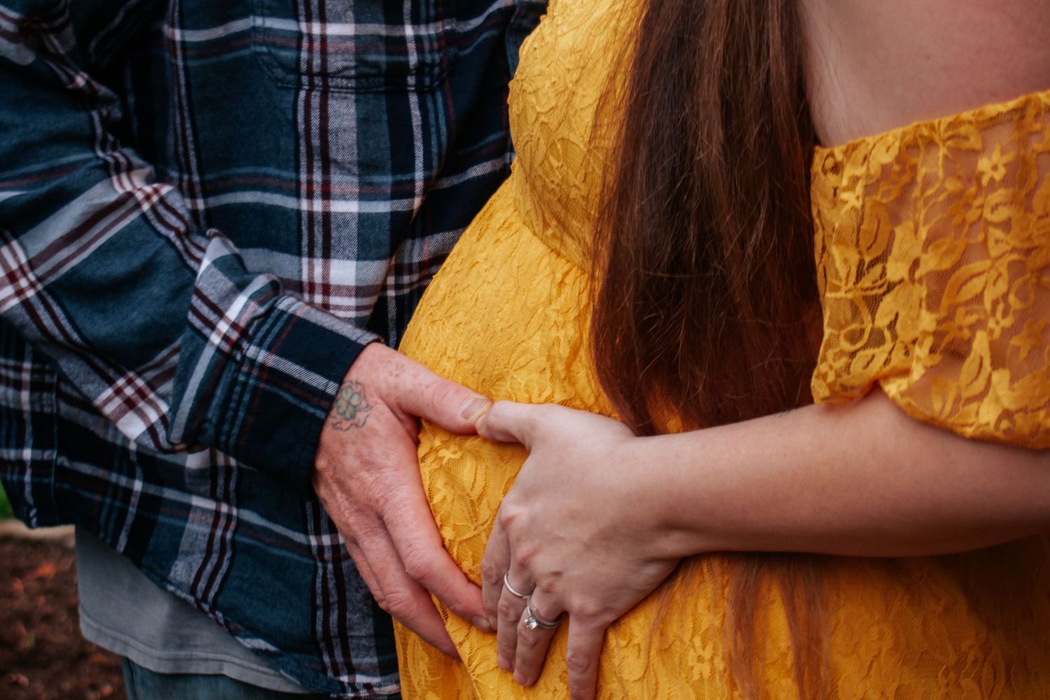 Couple holding pregnant belly together outdoors, woman in yellow lace dress with engagement ring.