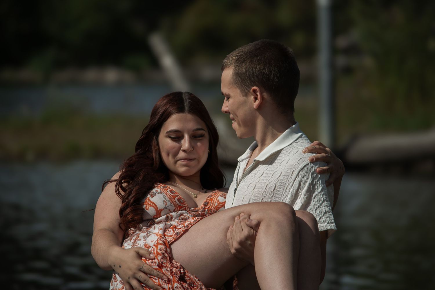 A man tenderly carries a pregnant woman in a floral dress near a serene lake backdrop.
