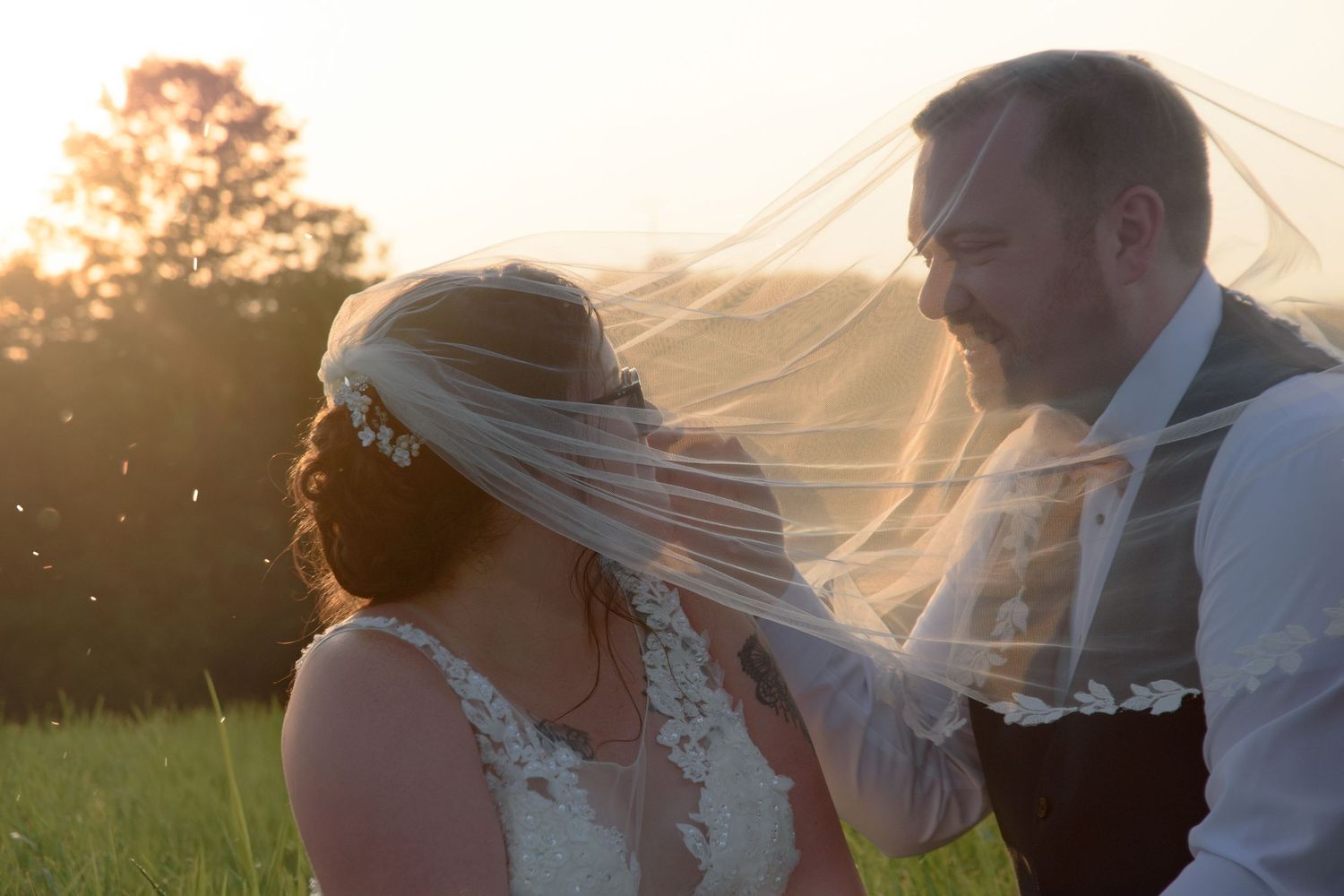 Bride and groom share a romantic sunset moment as veil billows in the breeze in a golden meadow.