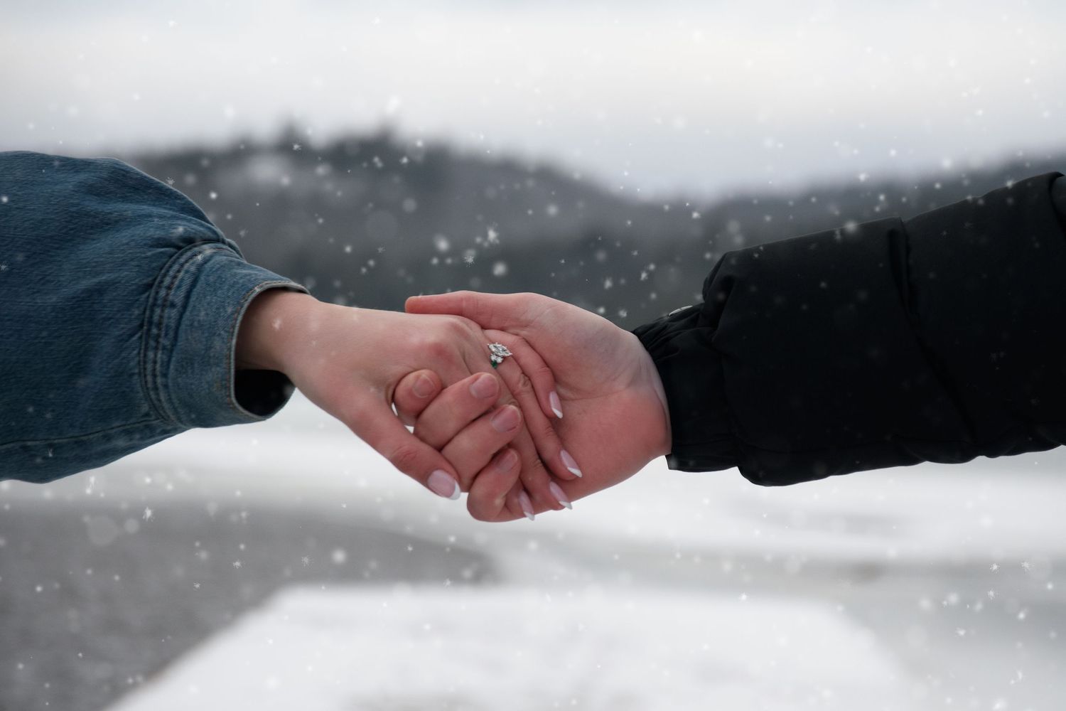 Couple holding hands with a diamond engagement ring visible while snow falls in a winter landscape.