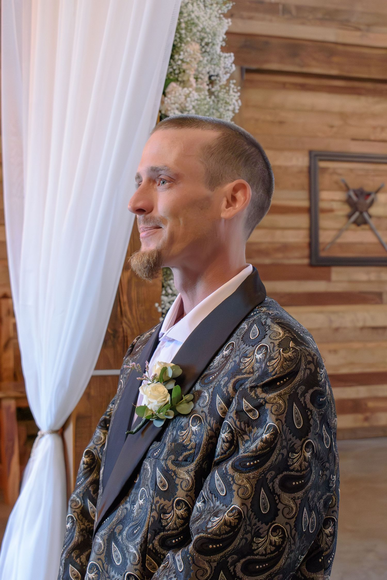 Smiling groom in paisley jacket with boutonniere stands at wedding altar with white drapes and wood wall.