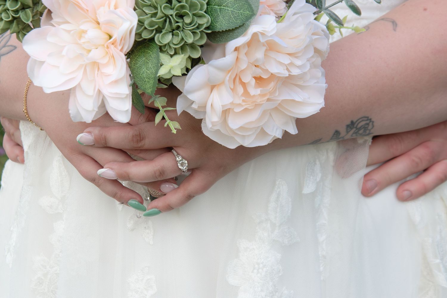Two brides holding hands with engagement ring, wearing white lace wedding dresses with tattoos and bouquet.