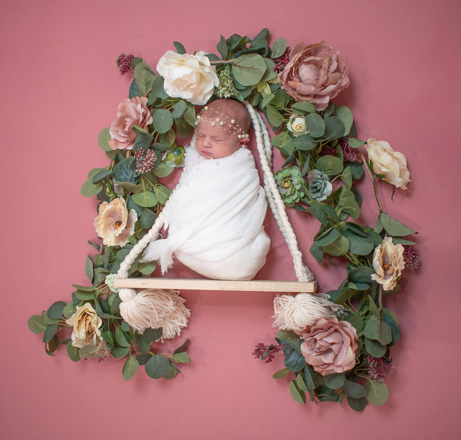 Sleeping newborn baby swaddled in white wrap on decorative swing surrounded by flowers.