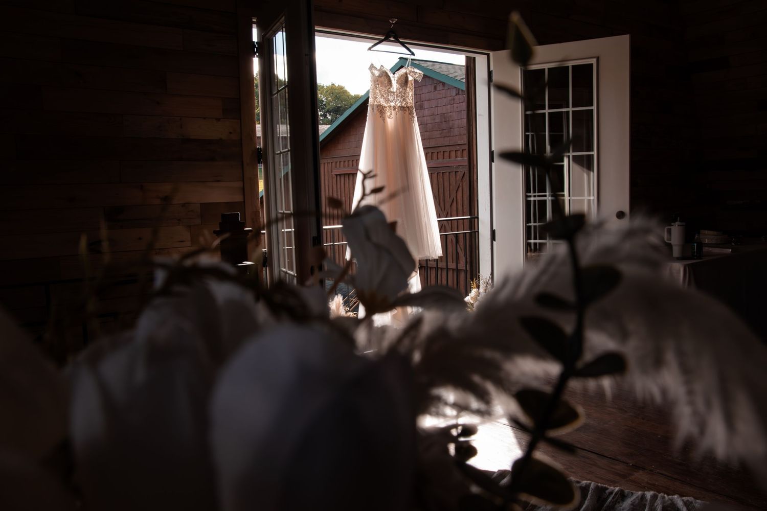 Wedding dress hanging in doorway of rustic barn venue, backlit by natural sunlight with floral details in foreground.