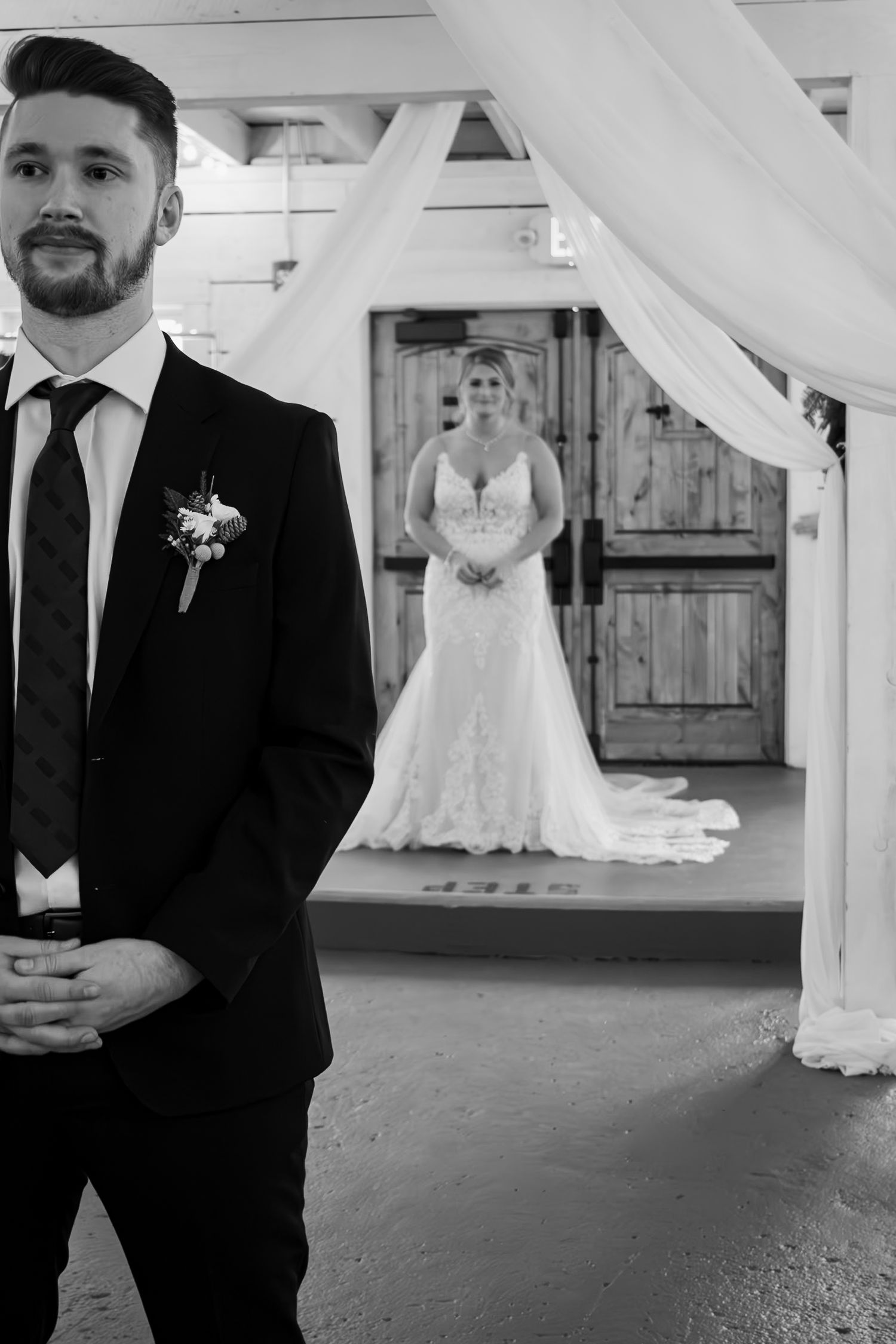 Black and white photo of a groom in a suit standing in foreground as bride in lace wedding dress waits behind him.