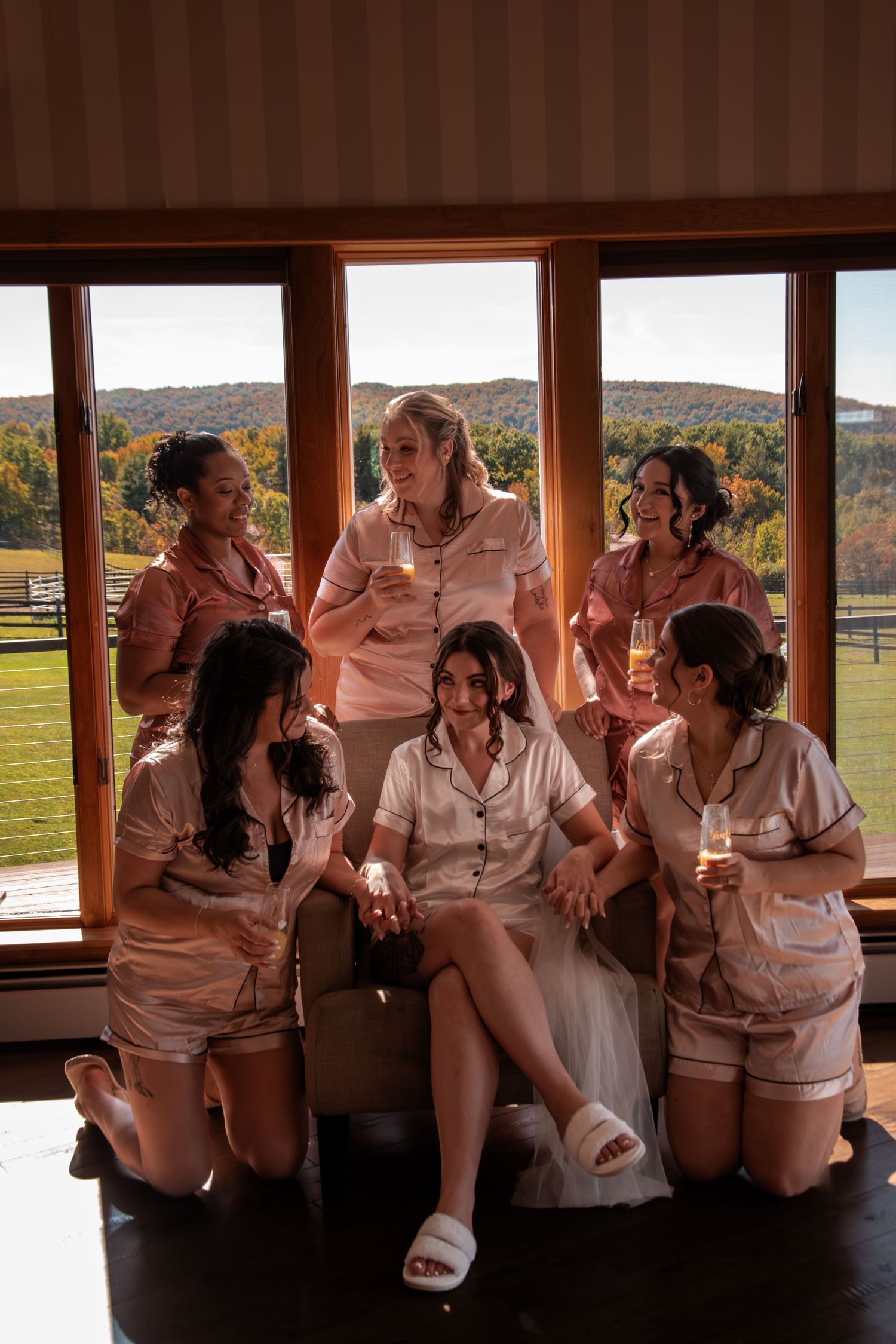 Bride and bridesmaids in matching satin pajamas celebrating together with champagne near scenic window.