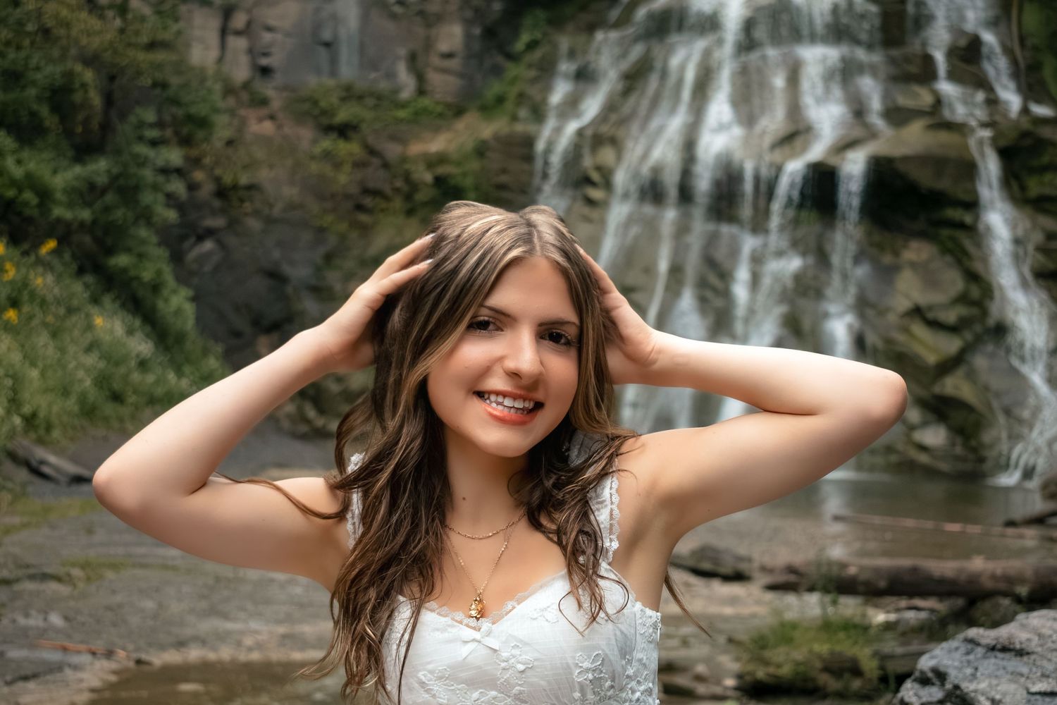 Young woman in white dress smiling with hands in hair, posing in front of a beautiful cascading waterfall.