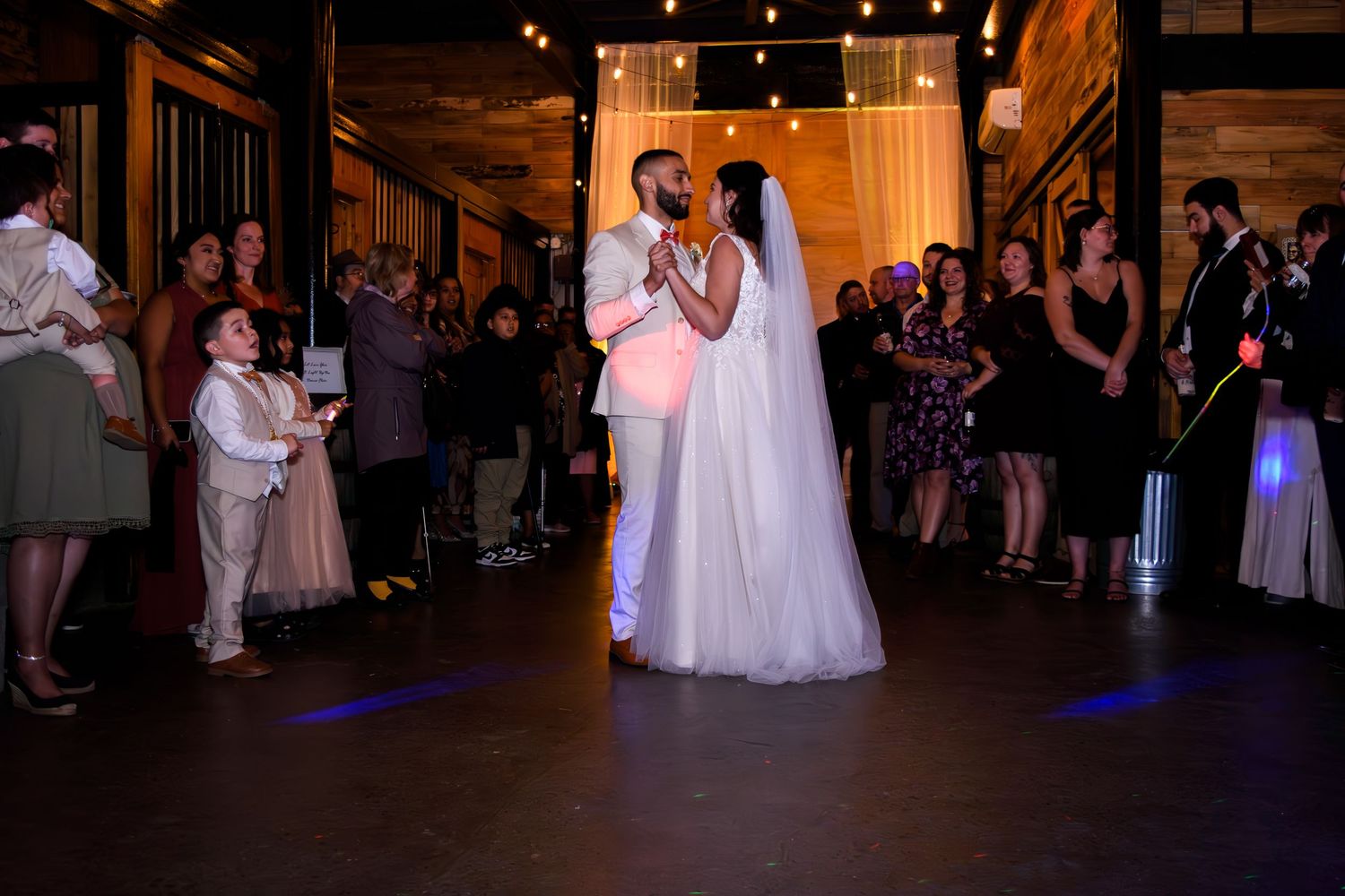 Bride and groom share their first dance at a wedding reception with guests watching in a warmly lit venue.