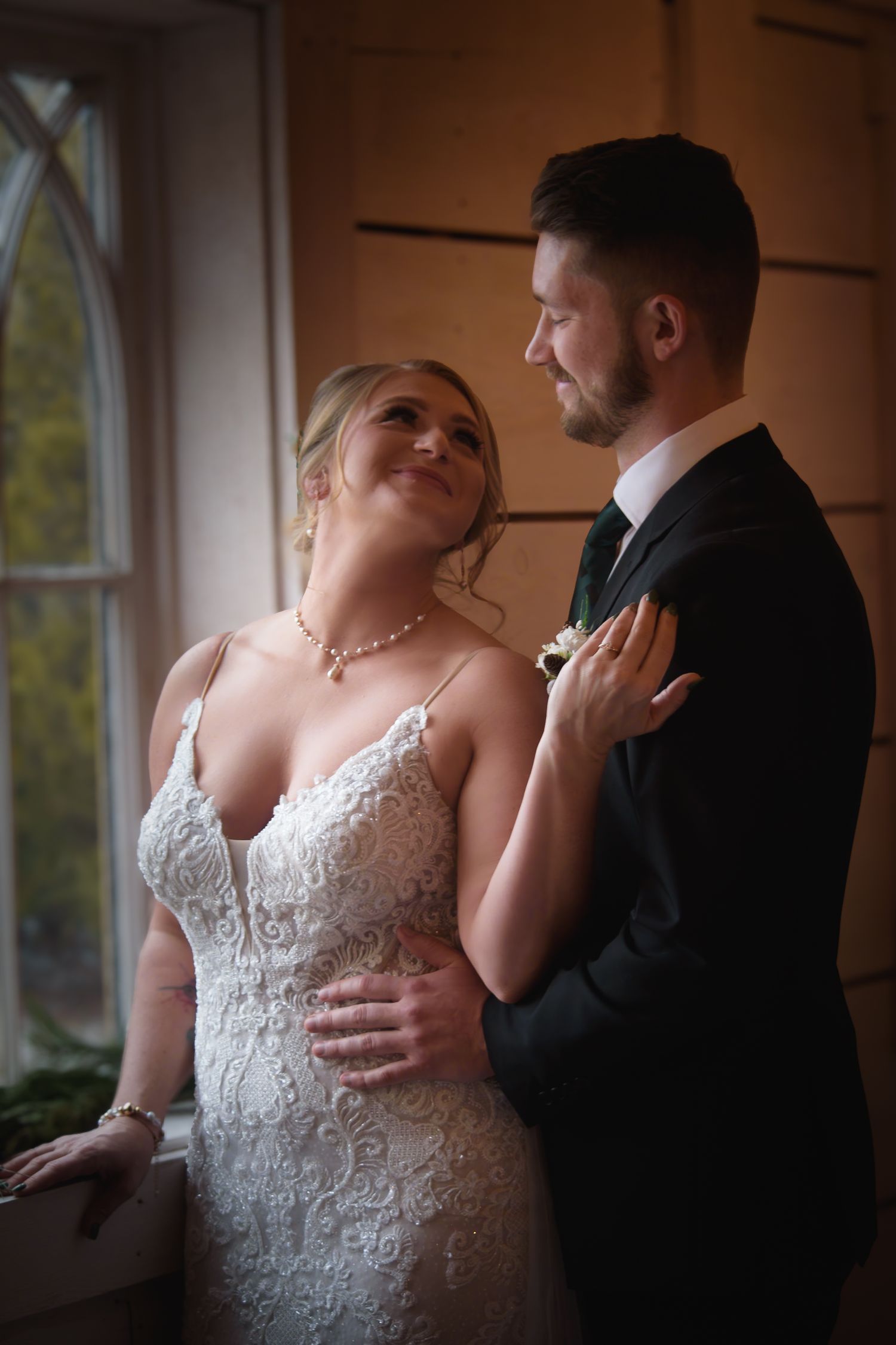 Bride in lace gown and groom in black suit sharing romantic moment by window.