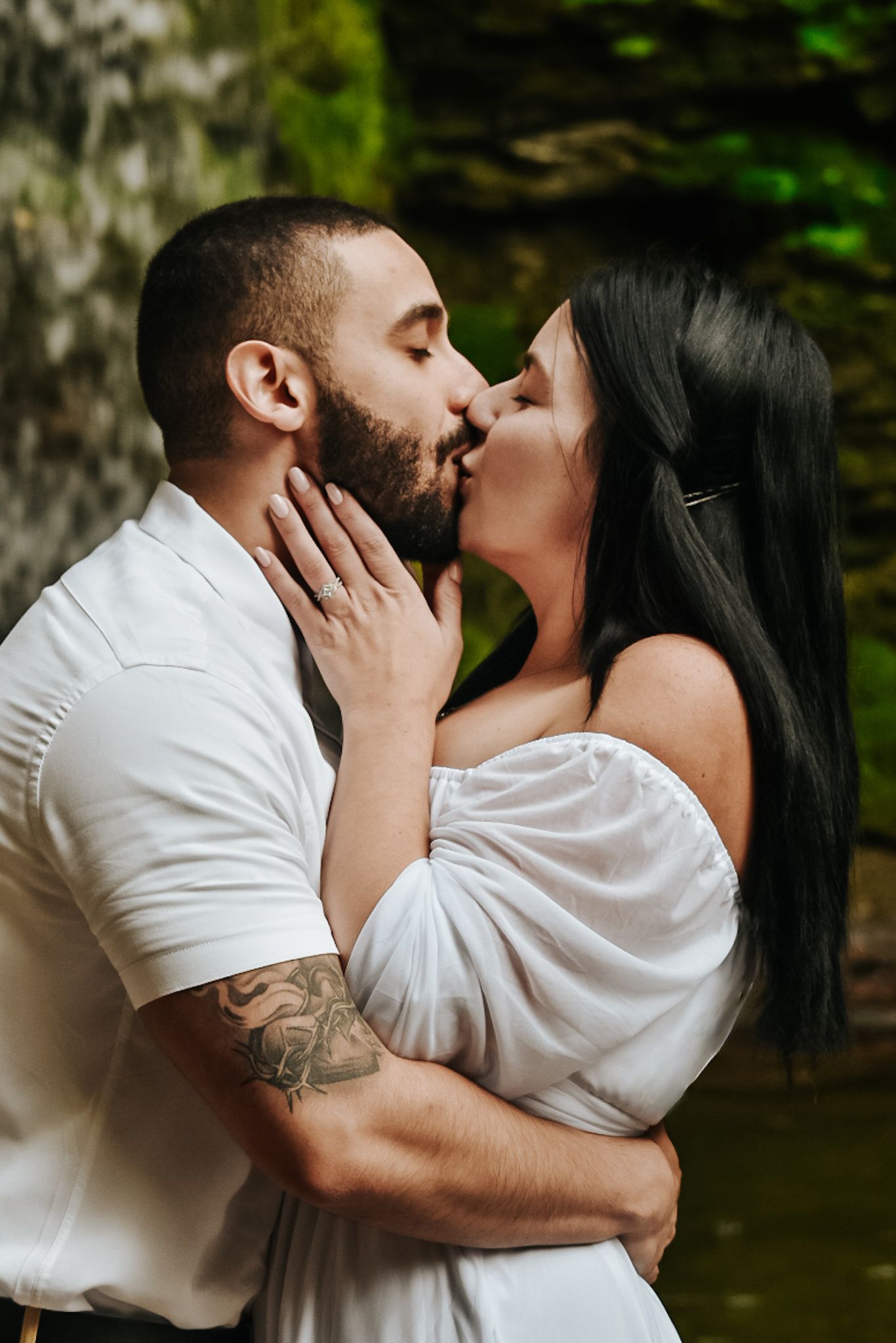 A bearded groom with tattoos kisses his dark-haired bride in a white off-shoulder dress outdoors.
