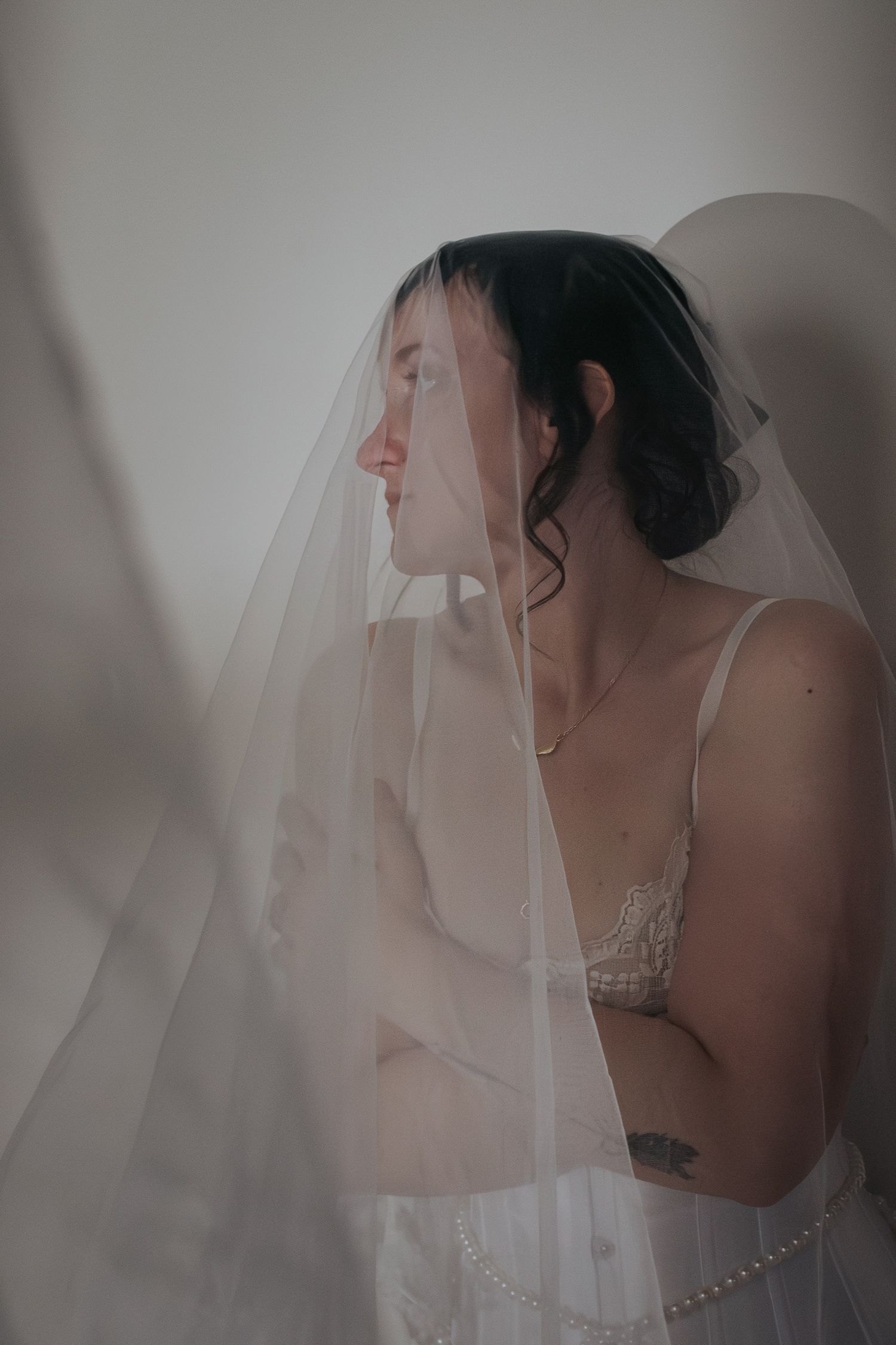 Bride in lace lingerie and sheer veil with arms crossed, soft light, tattoo visible, elegant bridal boudoir portrait.