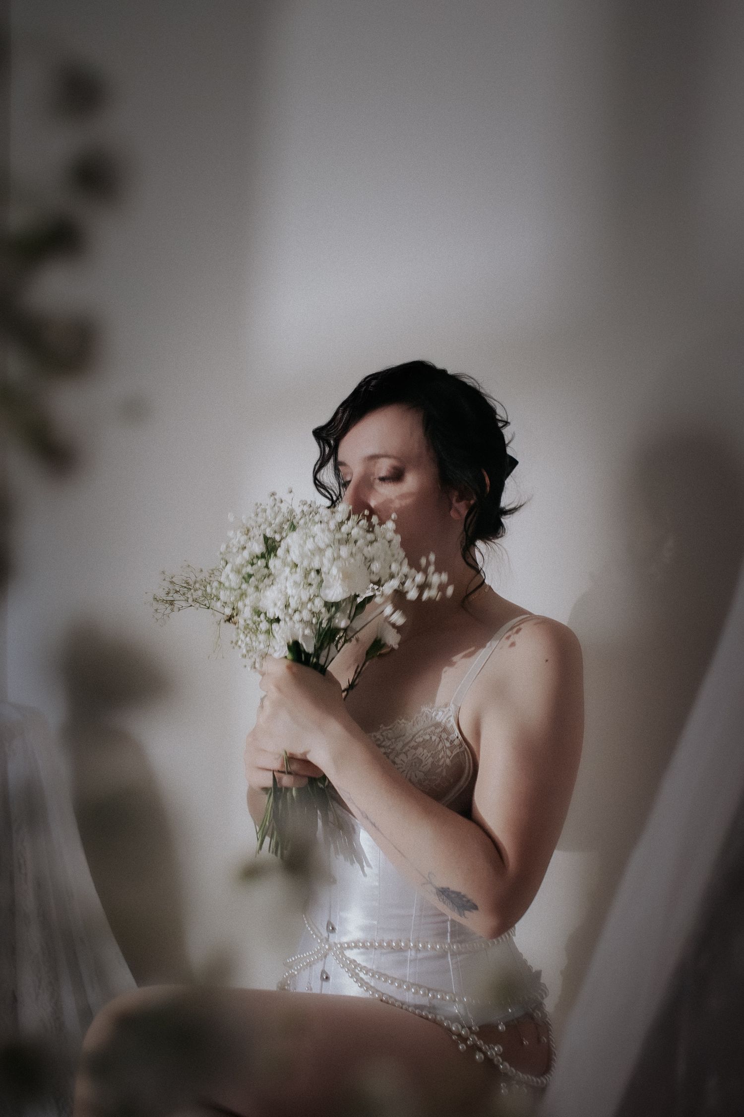 Bride in white corset smelling a delicate bouquet of white flowers and baby's breath in a softly lit room.