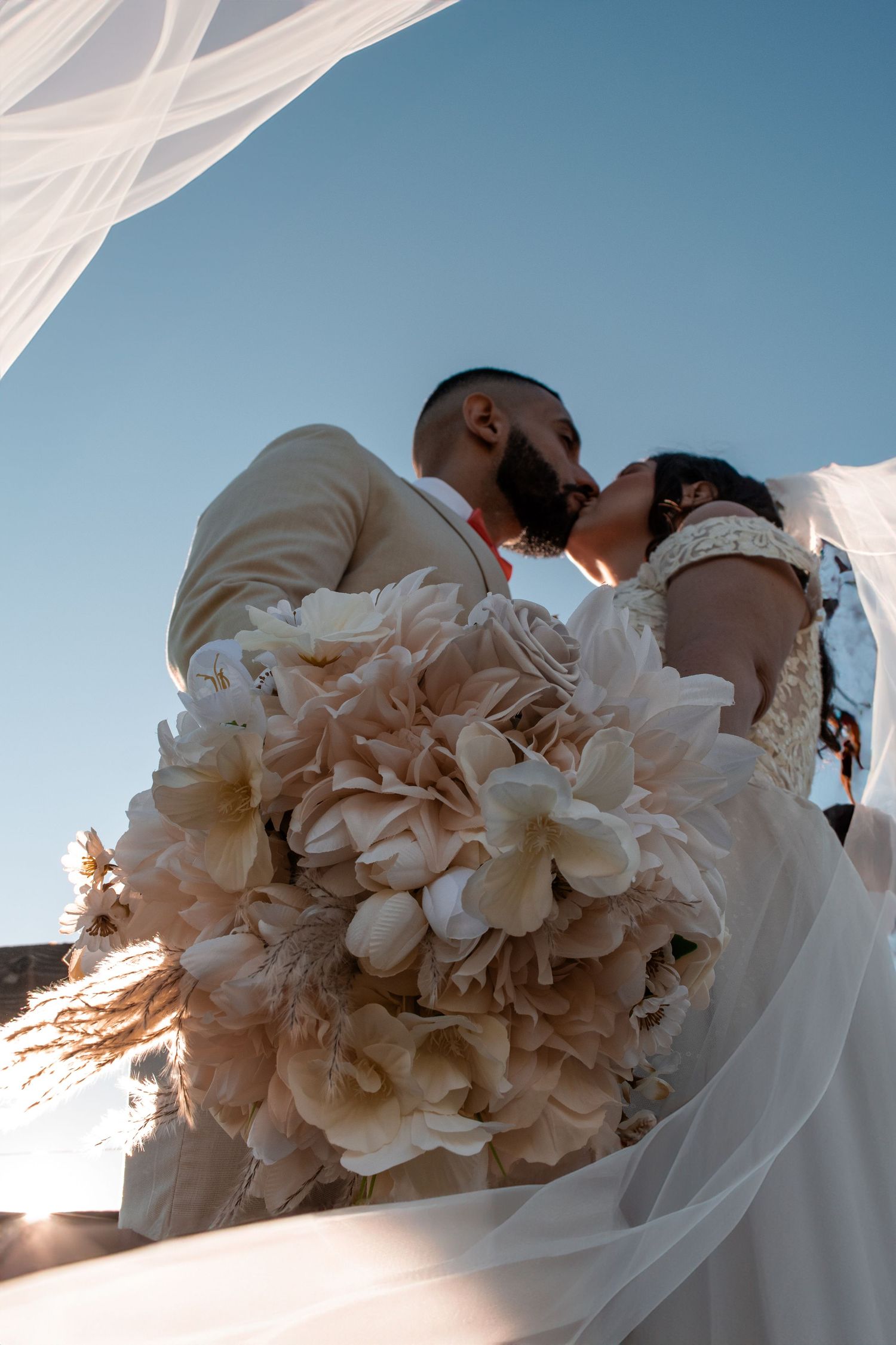 Bride and groom share a kiss outdoors, bride holding large blush floral bouquet, blue sky background.