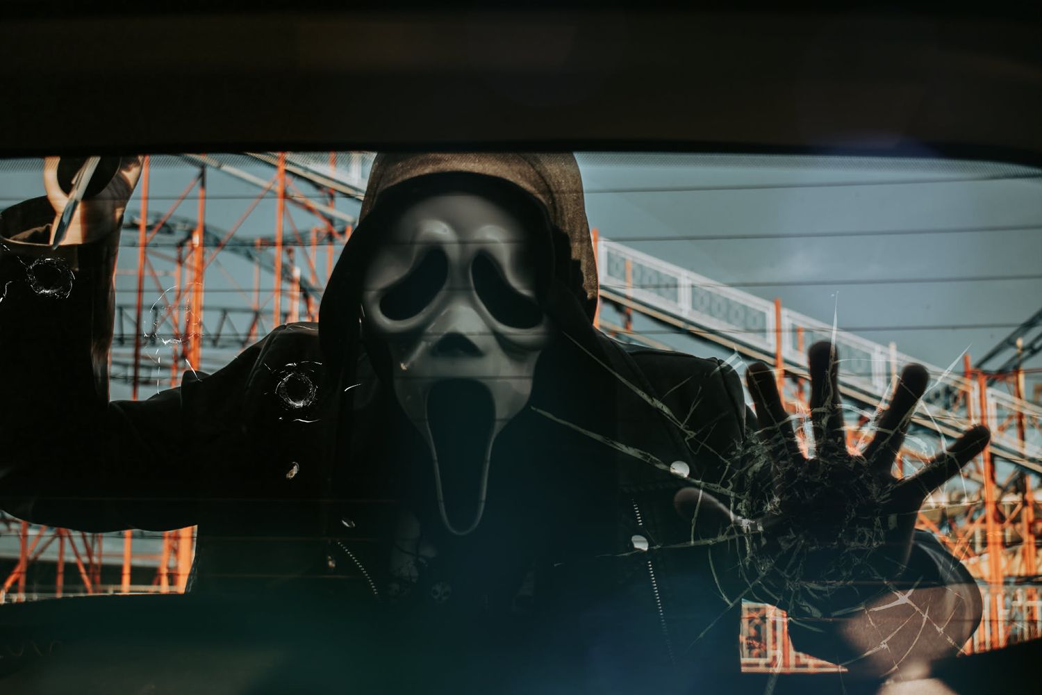 Person in Ghostface Halloween mask pressing hands against cracked glass in a dark, eerie urban setting.