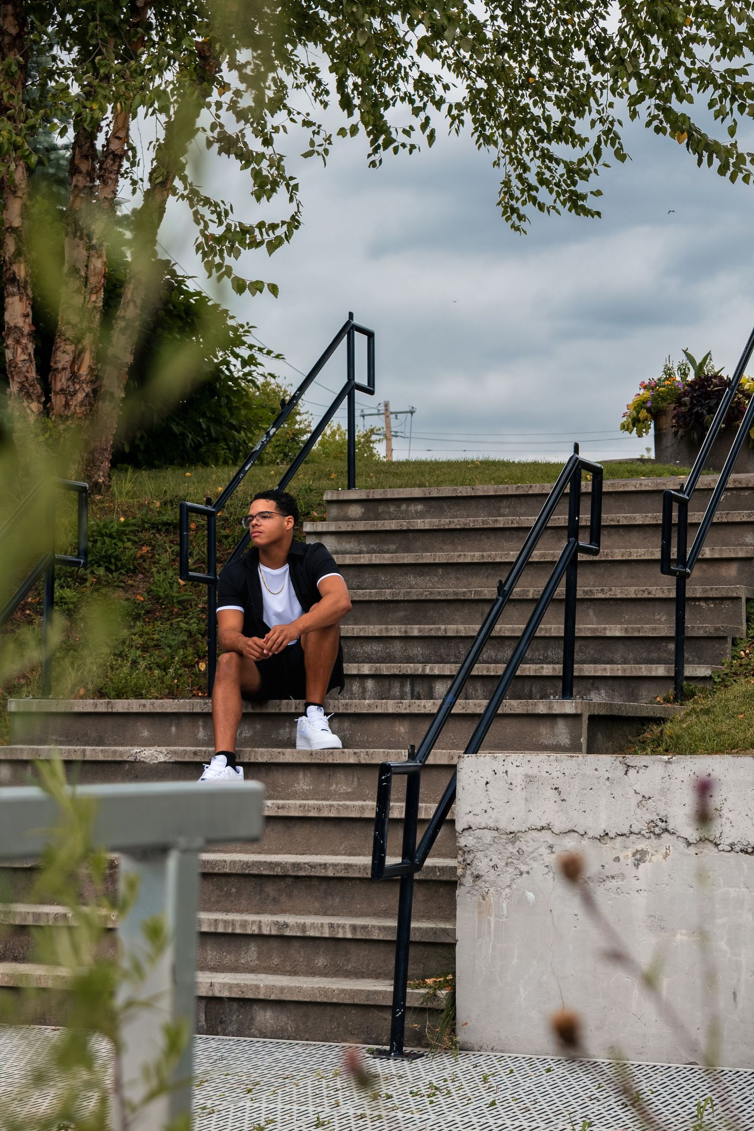 Young man in black shirt and white sneakers sitting on concrete steps outdoors, looking thoughtful under cloudy sky.