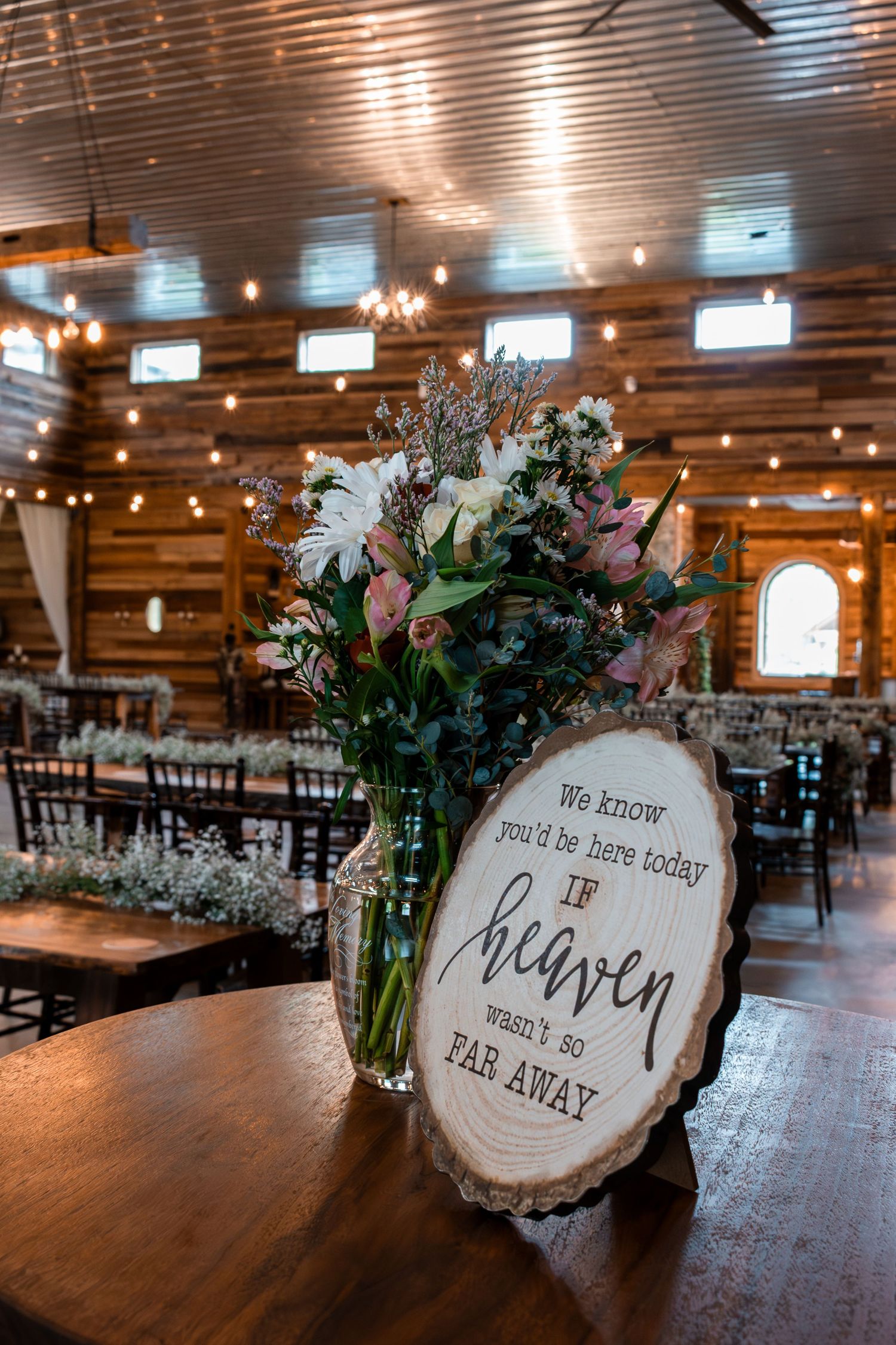 Rustic barn wedding venue with wood slice memorial sign reading heaven quote, floral arrangement, string lights, and wooden tables.