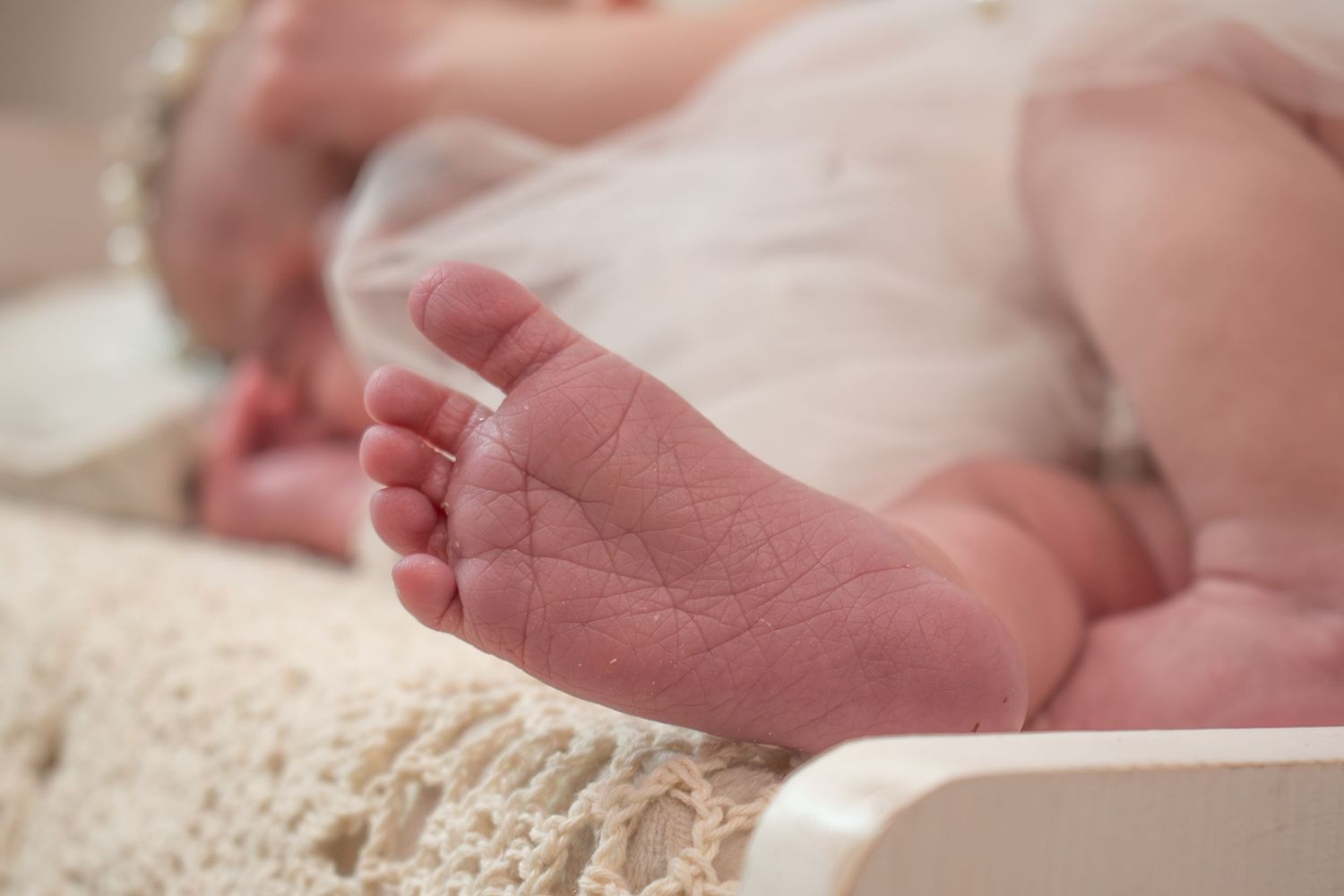 Close-up of tiny newborn baby feet resting on a soft white lace blanket in a delicate pink outfit.