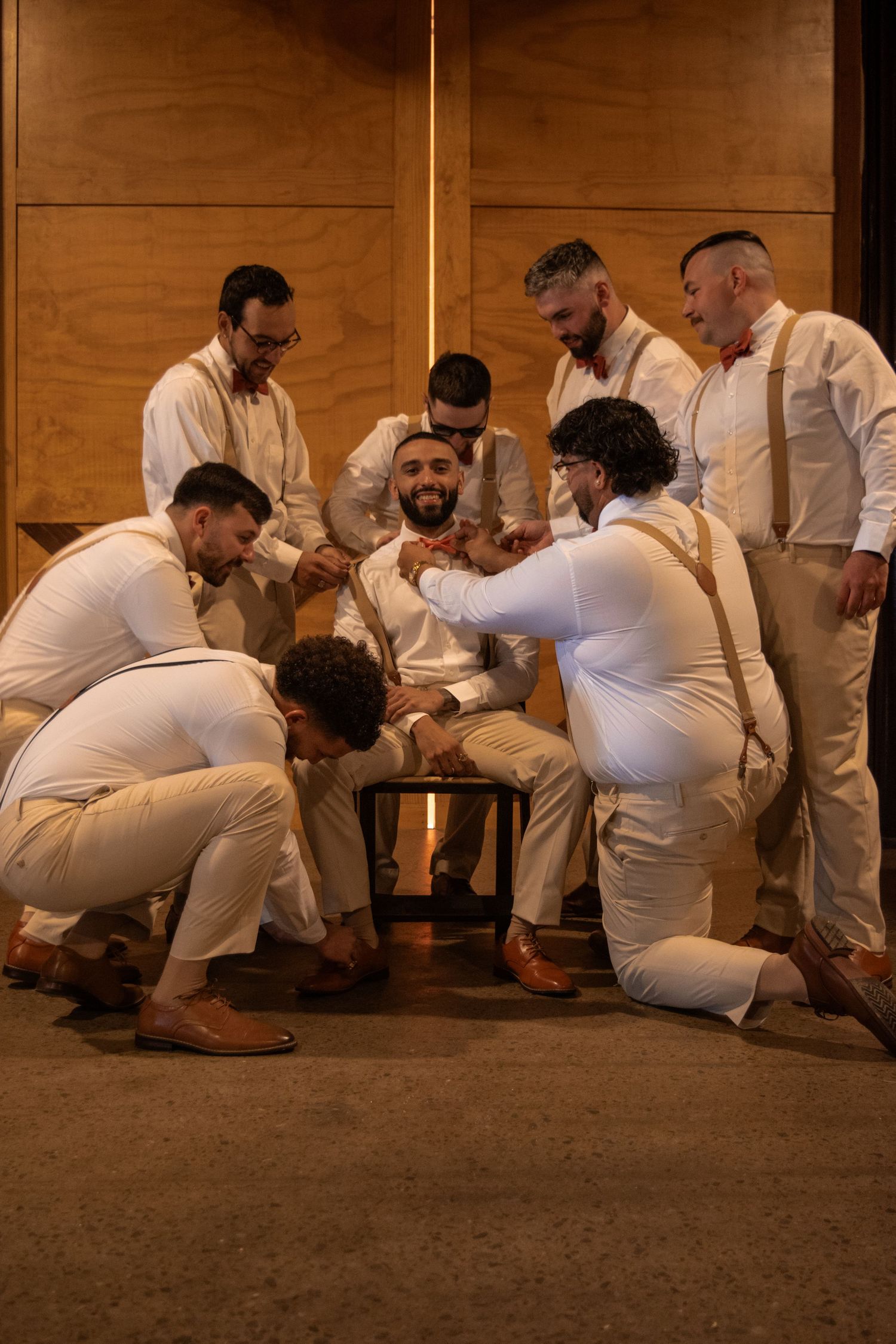 Groom surrounded by groomsmen in white suits helping him prepare in warm-lit wedding venue.