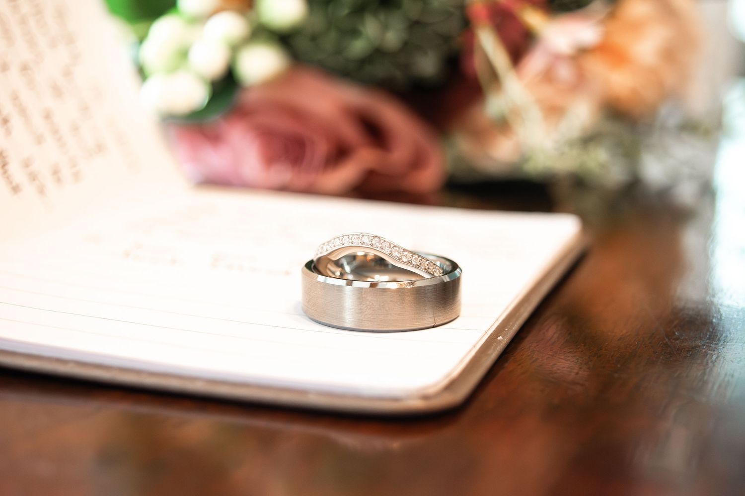 Two silver wedding rings resting on an open notebook with flowers in the background.