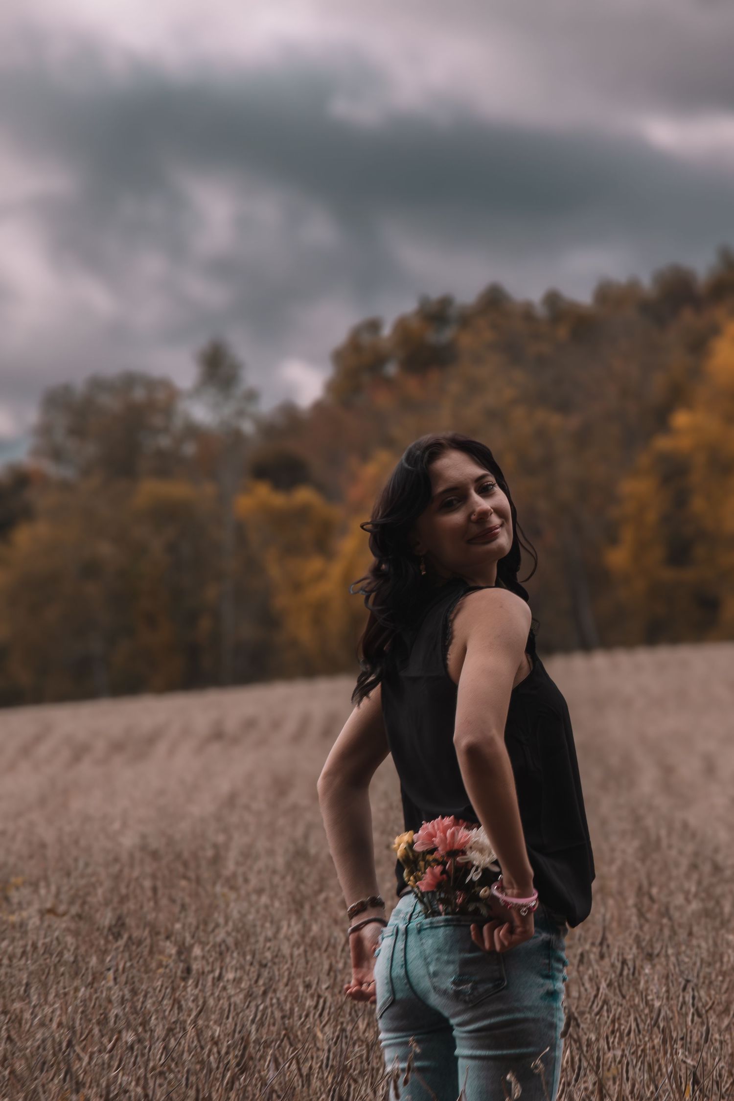 Young woman in black top holding flowers standing in an autumn field with cloudy sky and colorful trees.