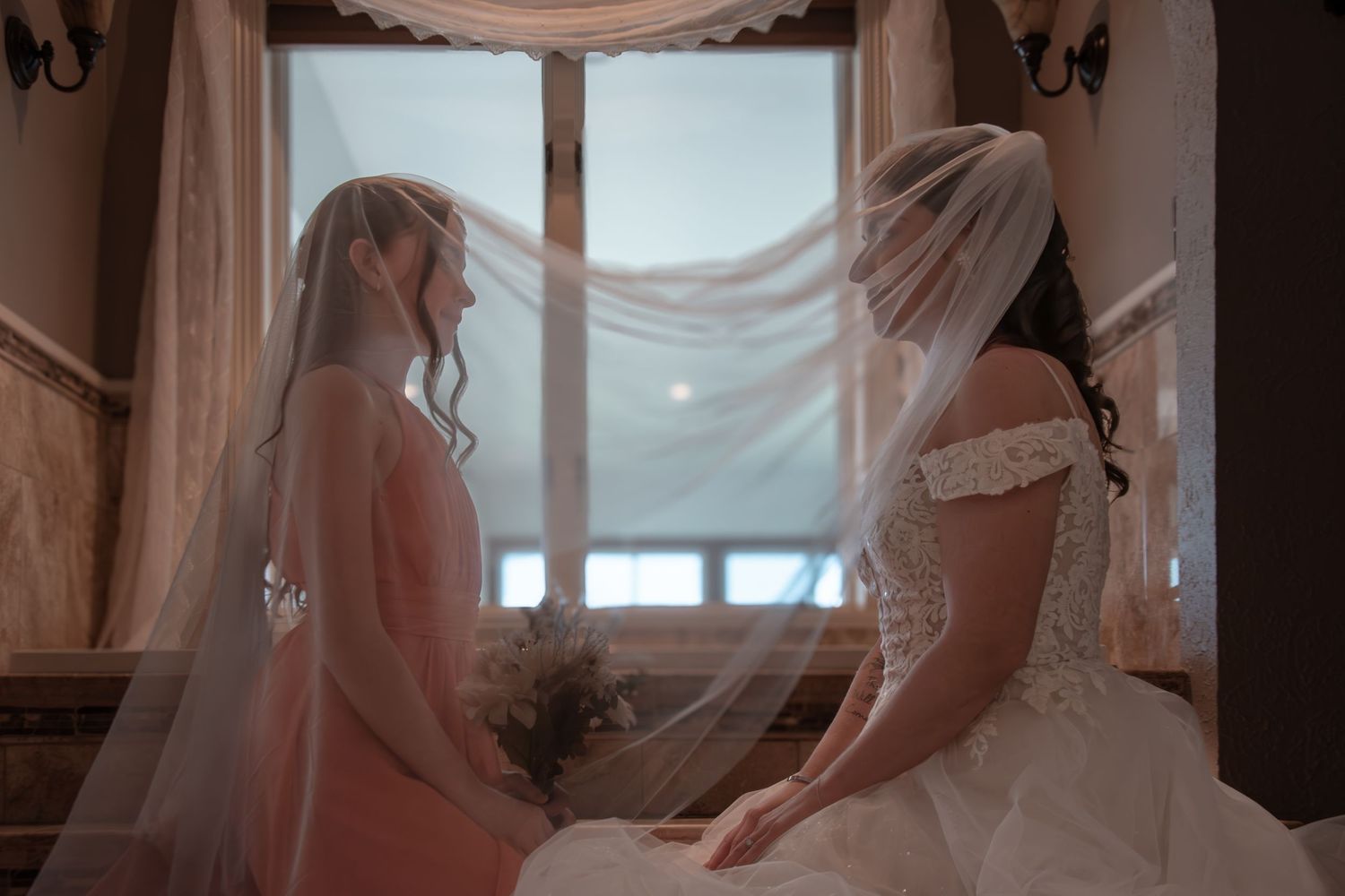 Two brides in wedding gowns and veils sit facing each other near a sunlit window in an elegant venue.
