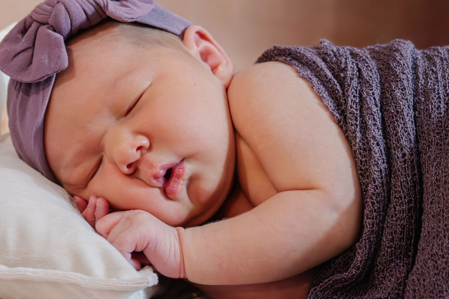 Sleeping newborn baby girl wearing purple bow headband wrapped in lavender swaddle on pillow.