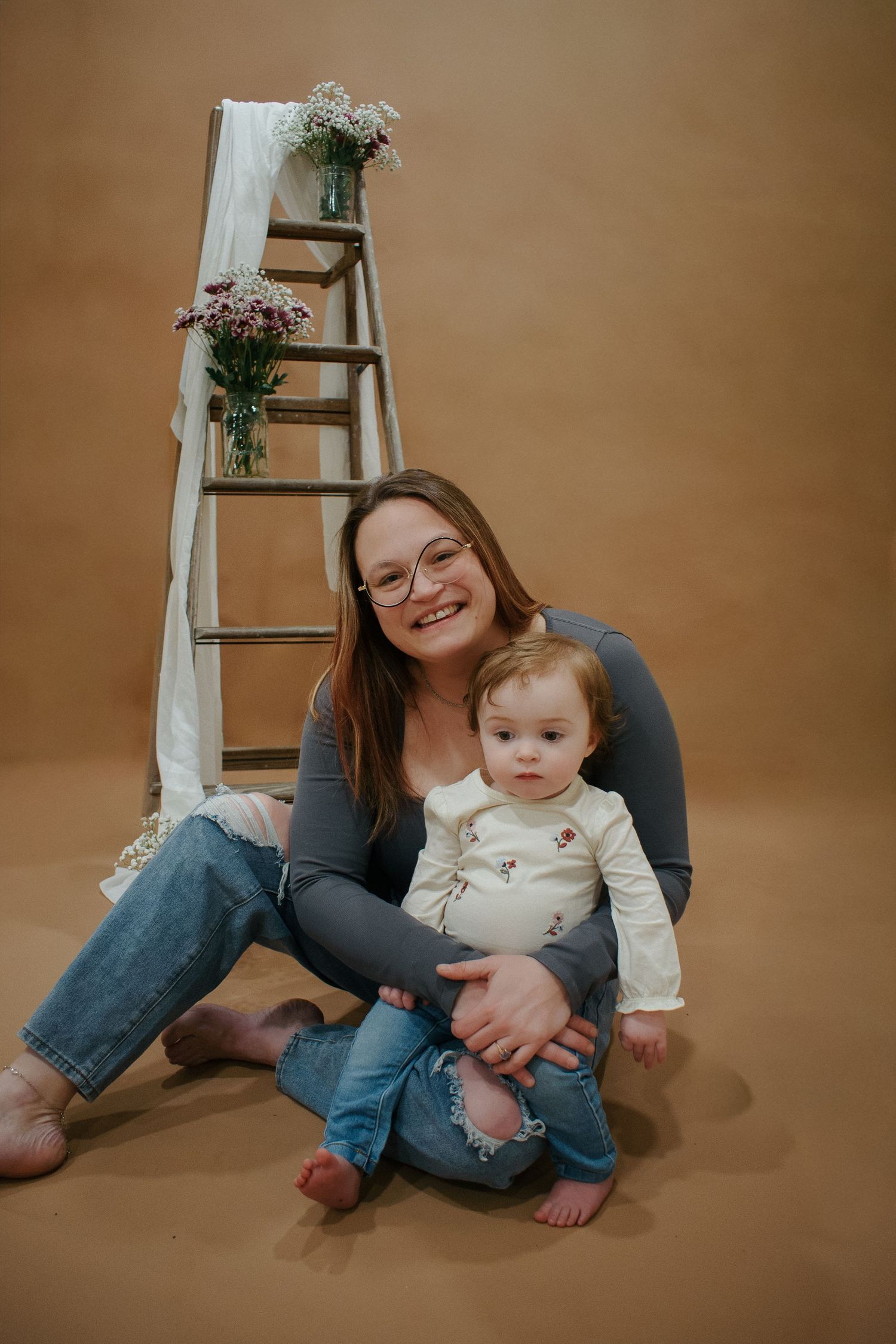 A smiling woman with glasses sits on the floor holding a baby, posed in front of a decorated wooden ladder with flowers.