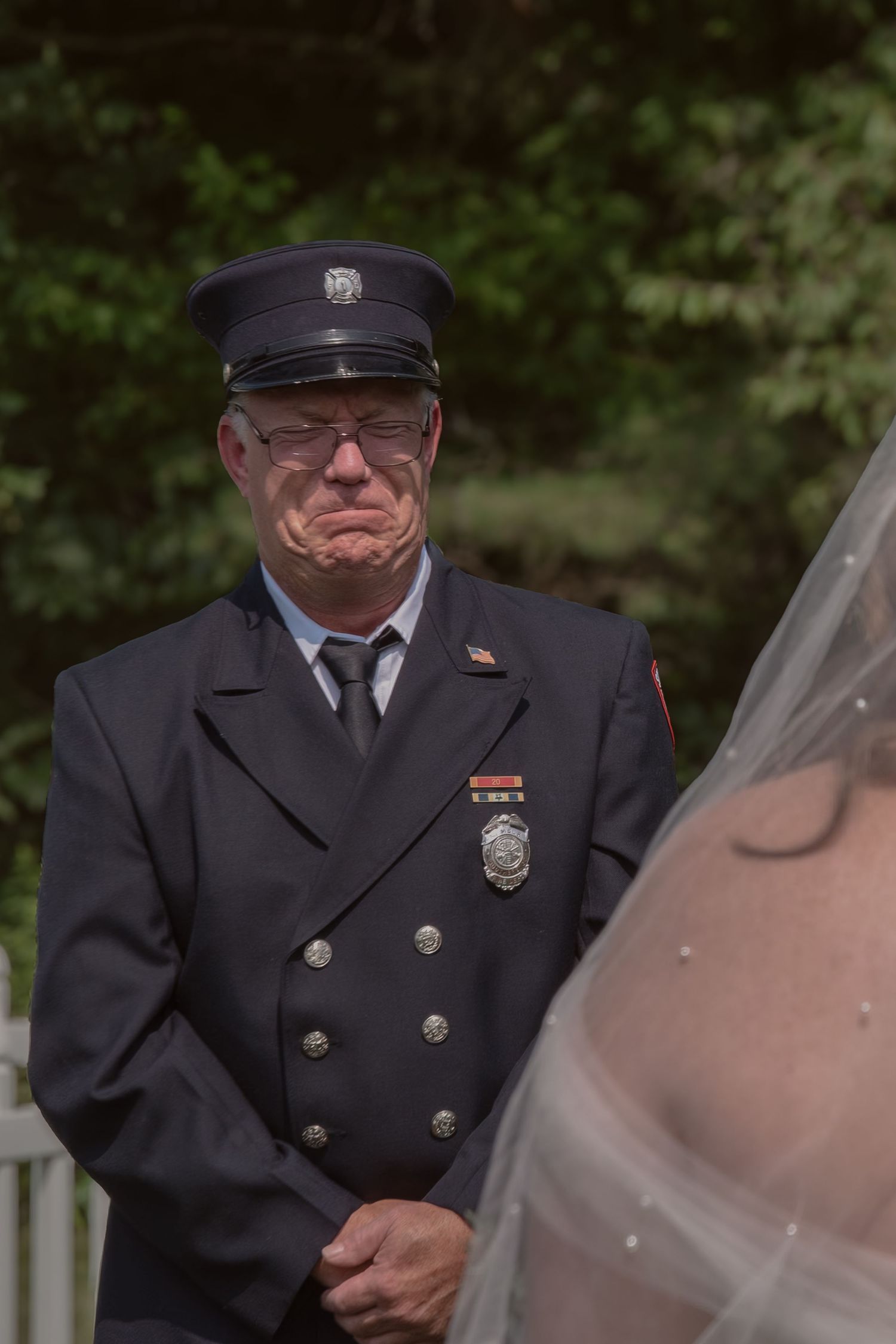 Emotional firefighter in dress uniform watching bride during outdoor wedding ceremony.