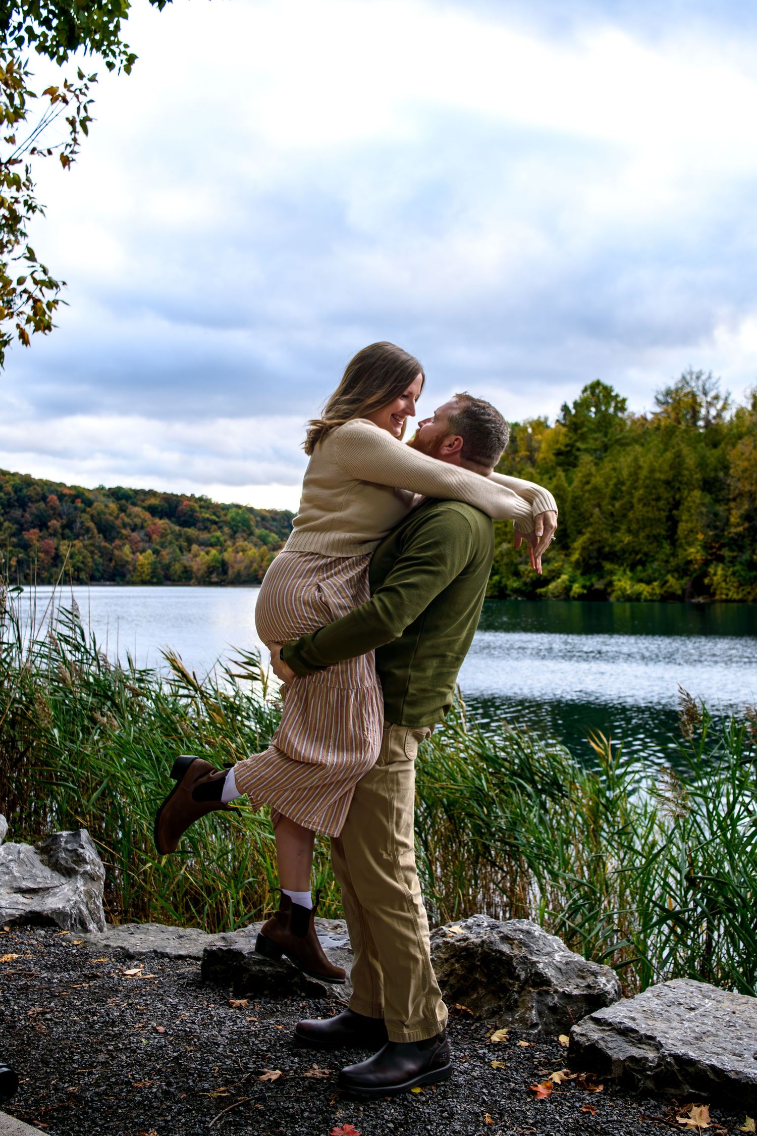 Couple embracing and laughing by a scenic lake surrounded by autumn foliage.