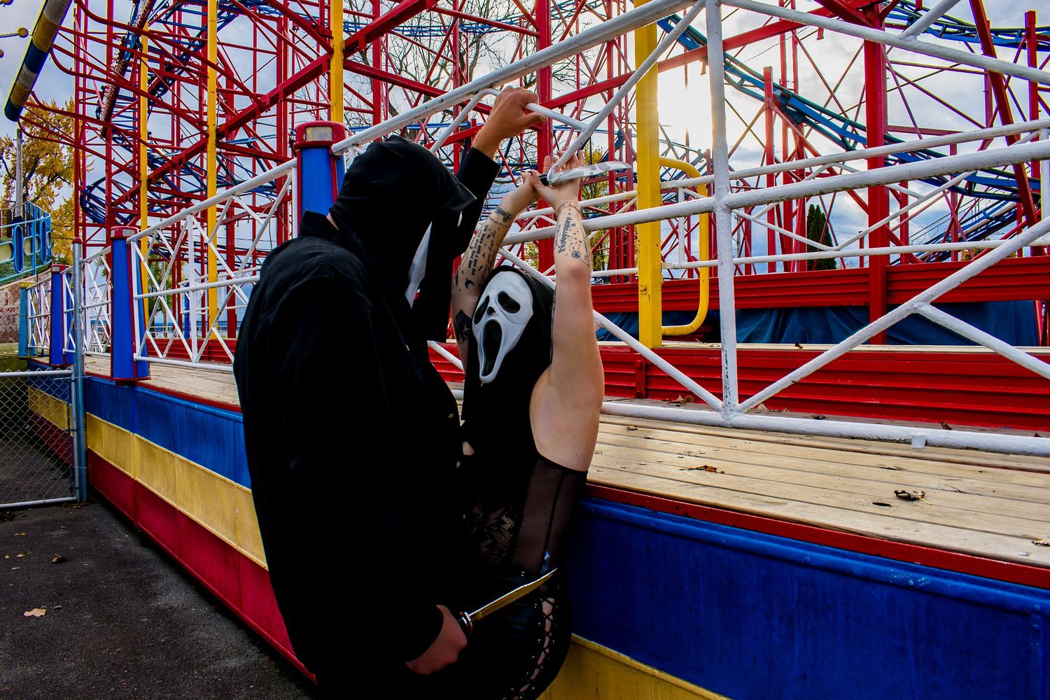 Couple in Ghostface Halloween costumes embracing near colorful carnival roller coaster ride.