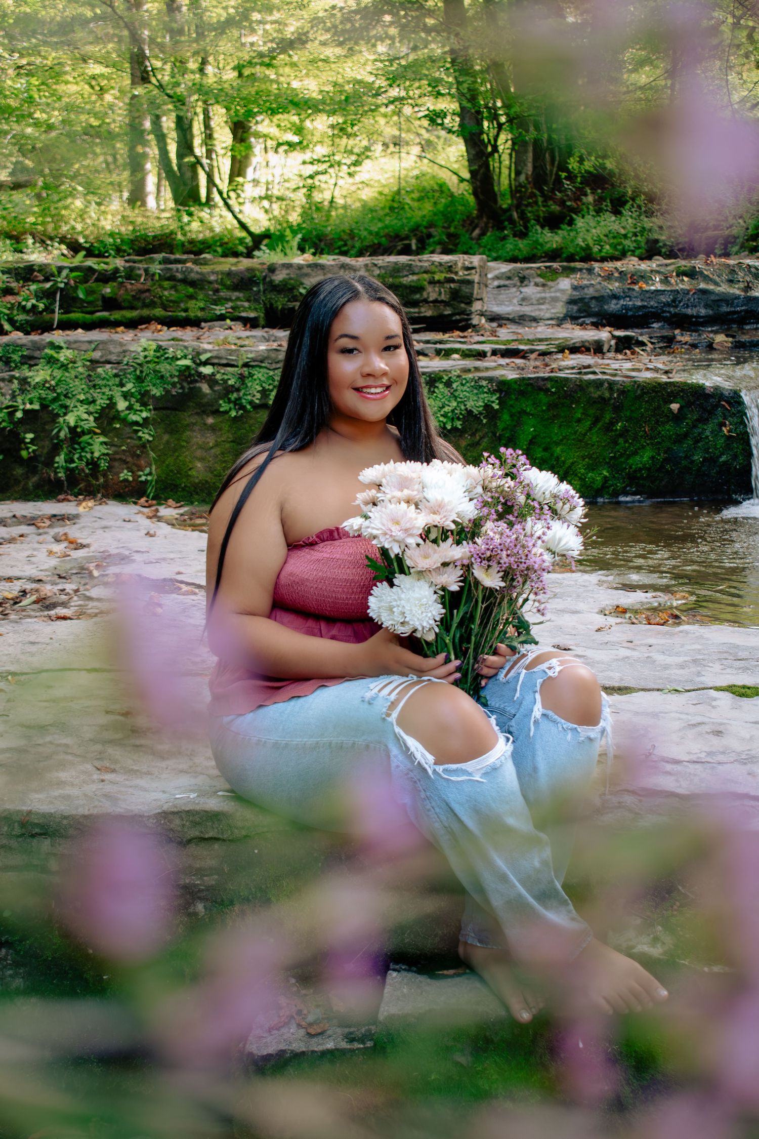 Young woman sitting on rocks by a stream holding white and pink flowers, wearing ripped jeans and a pink top.