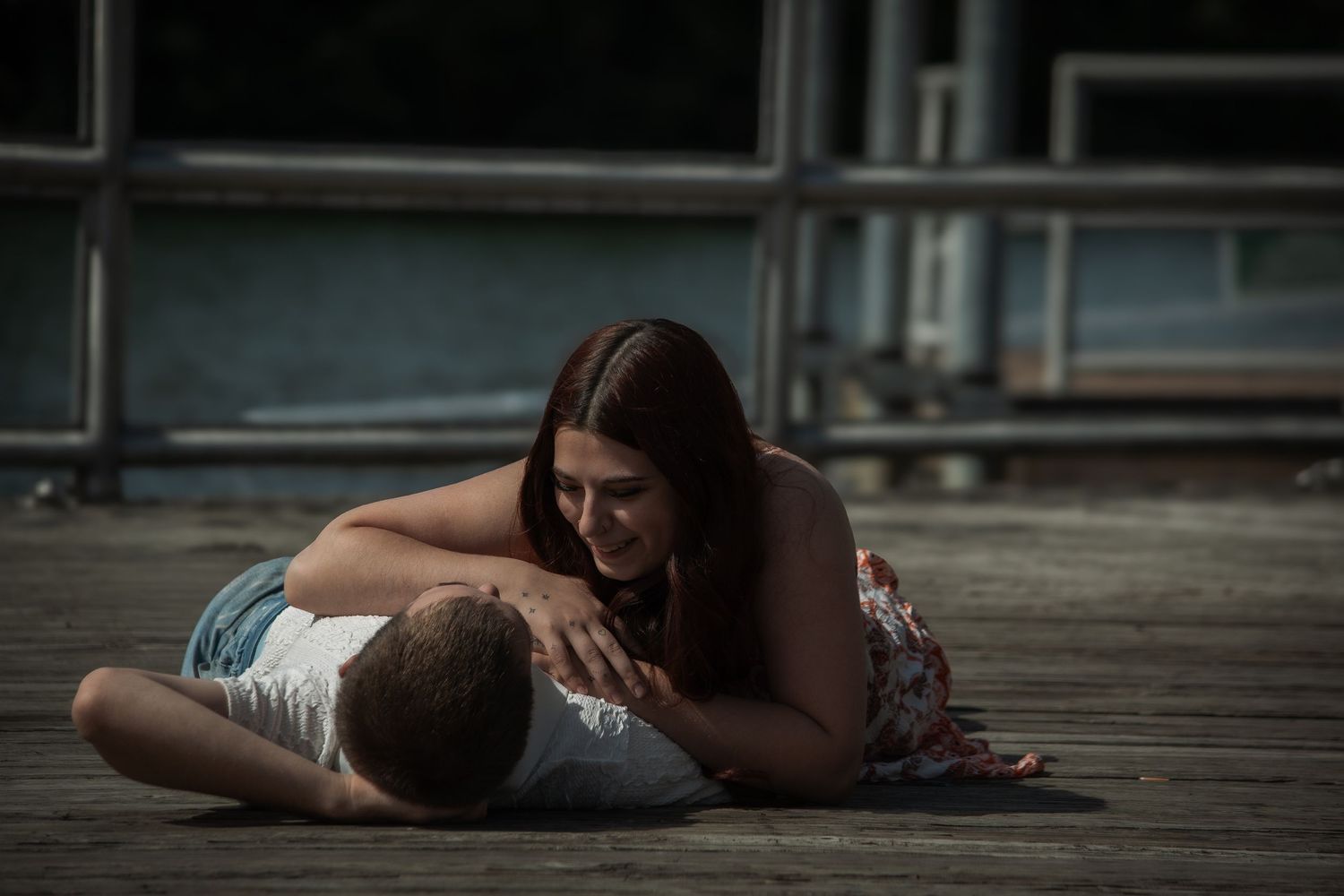A young woman cradles a man lying on a wooden dock in an emotional and intimate outdoor scene.