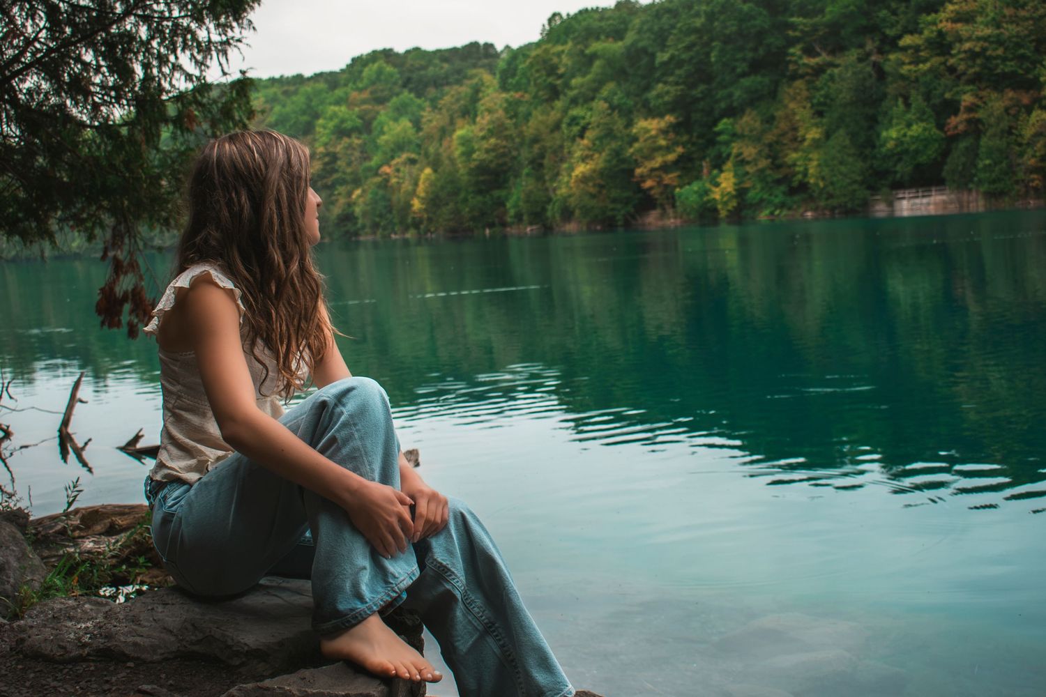 A young woman sits barefoot on a rock by a serene turquoise lake surrounded by lush green forest trees.