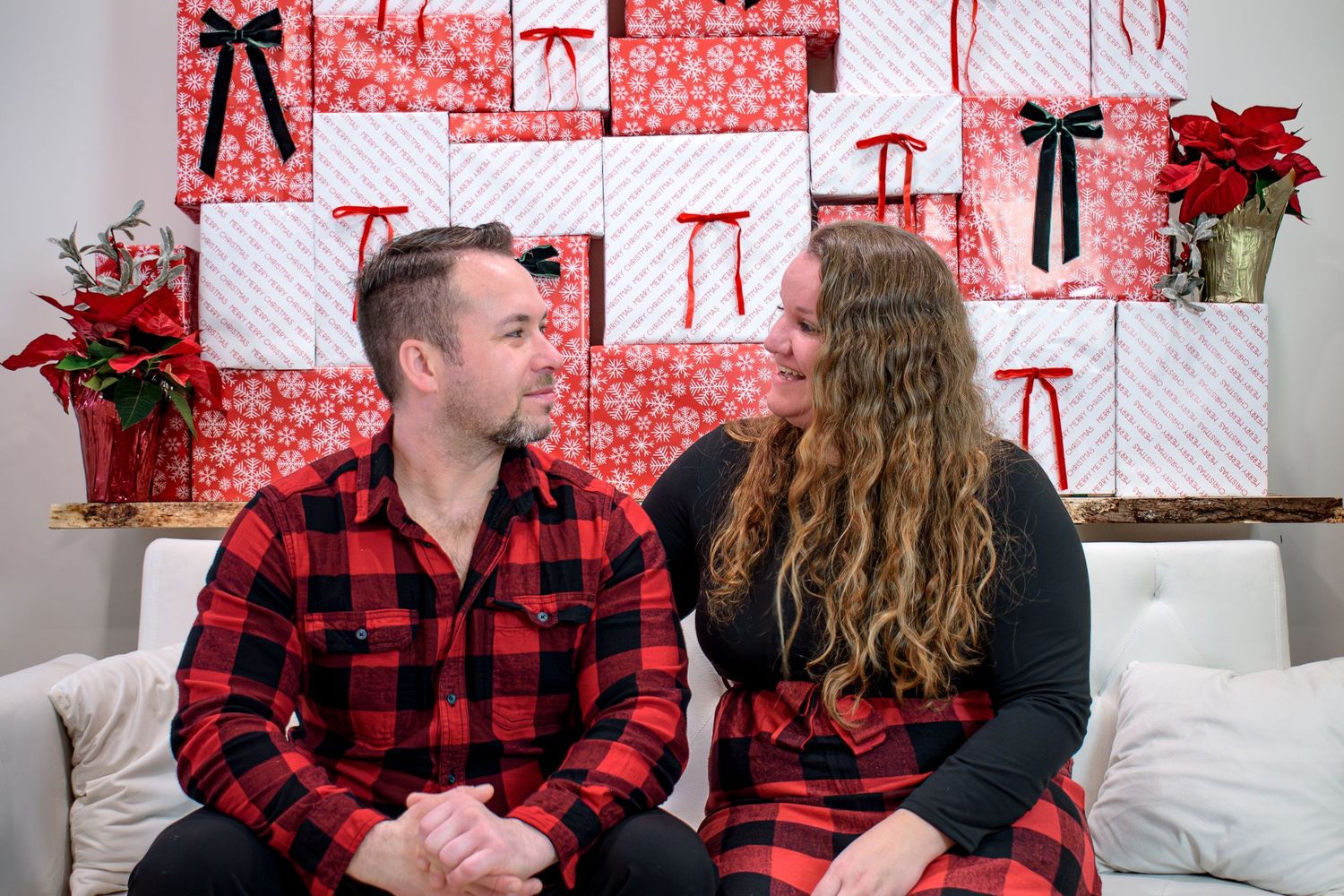 Couple in matching red plaid pajamas sitting on a couch in front of a Christmas gift wall.