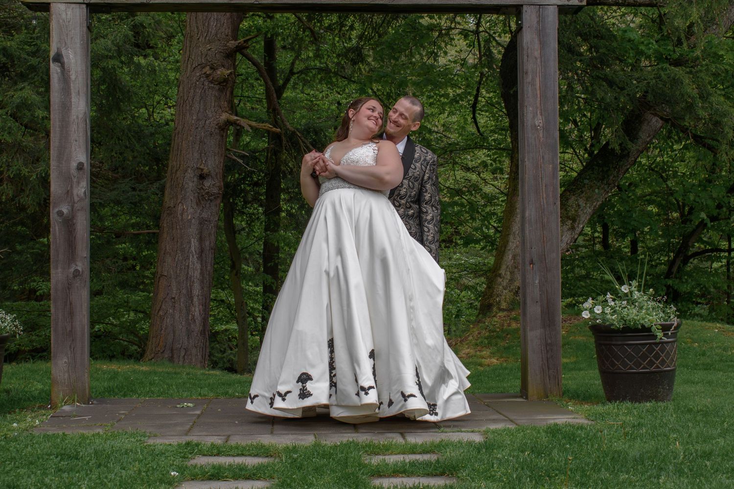 Couple embracing under wooden arbor in lush garden, bride in white gown with black accents, outdoor wedding ceremony.