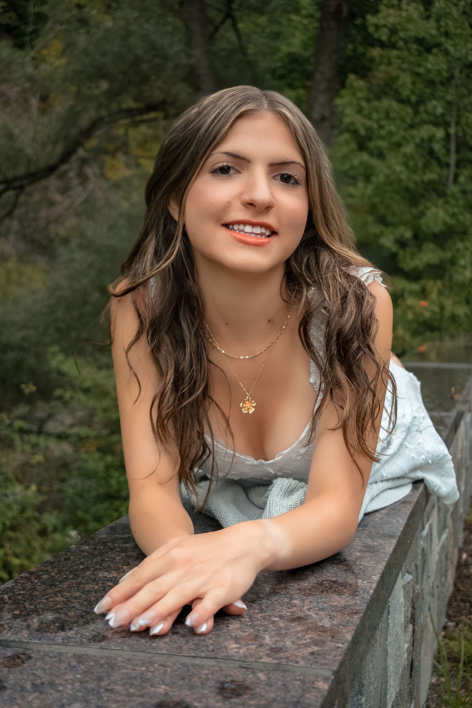Young woman with long wavy hair smiles while leaning on a stone railing outdoors, wearing a gold necklace.