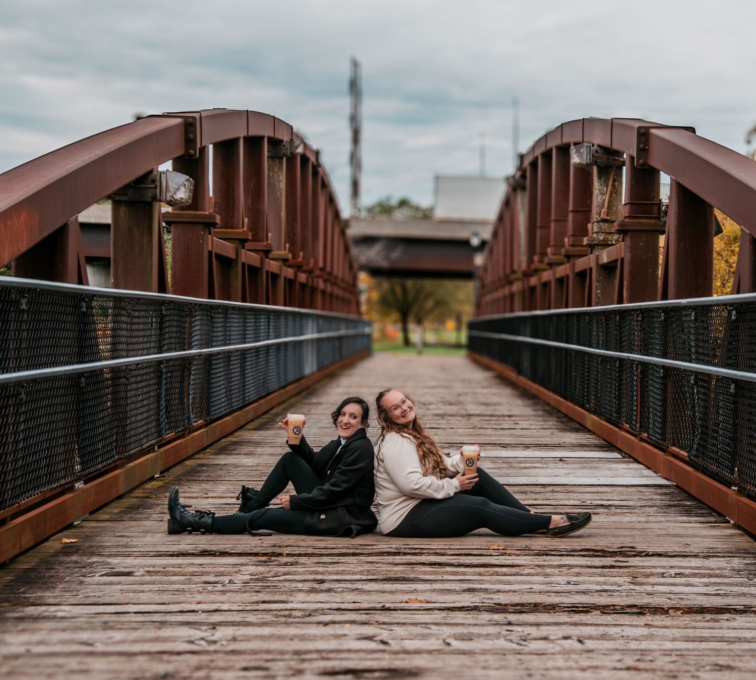Two friends sitting back-to-back on a rustic wooden bridge, smiling and posing for a photo on a cloudy day.