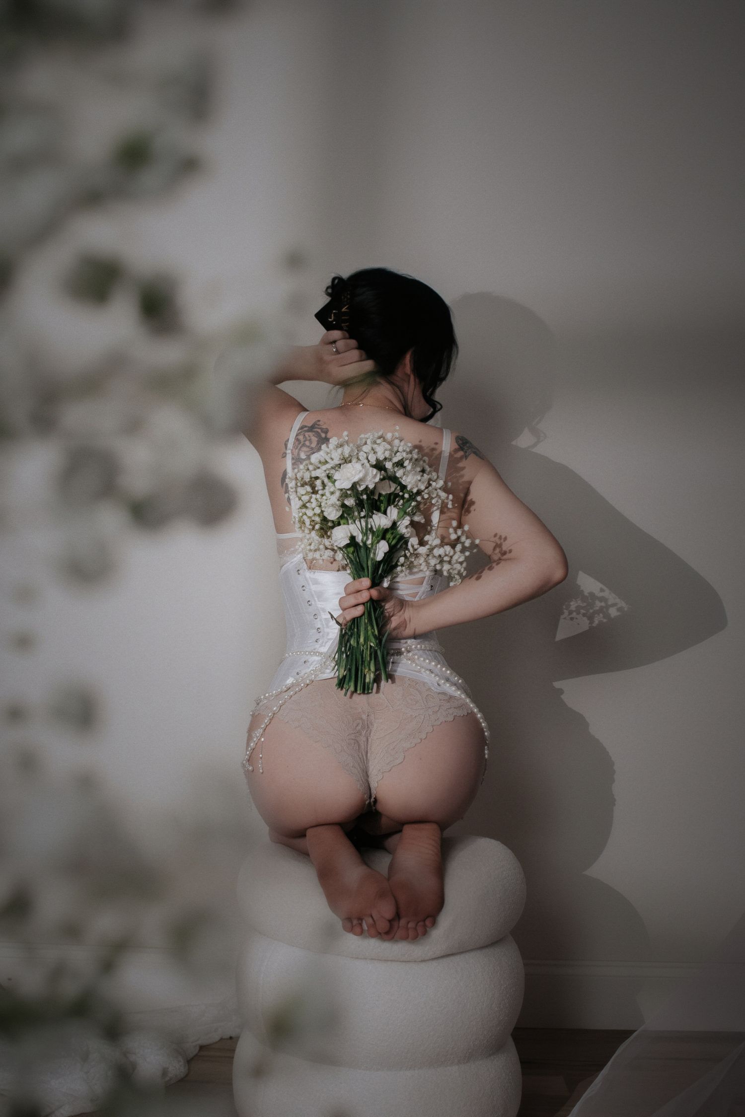 Woman in white lace lingerie holding a bouquet of white flowers, posed against a light wall.