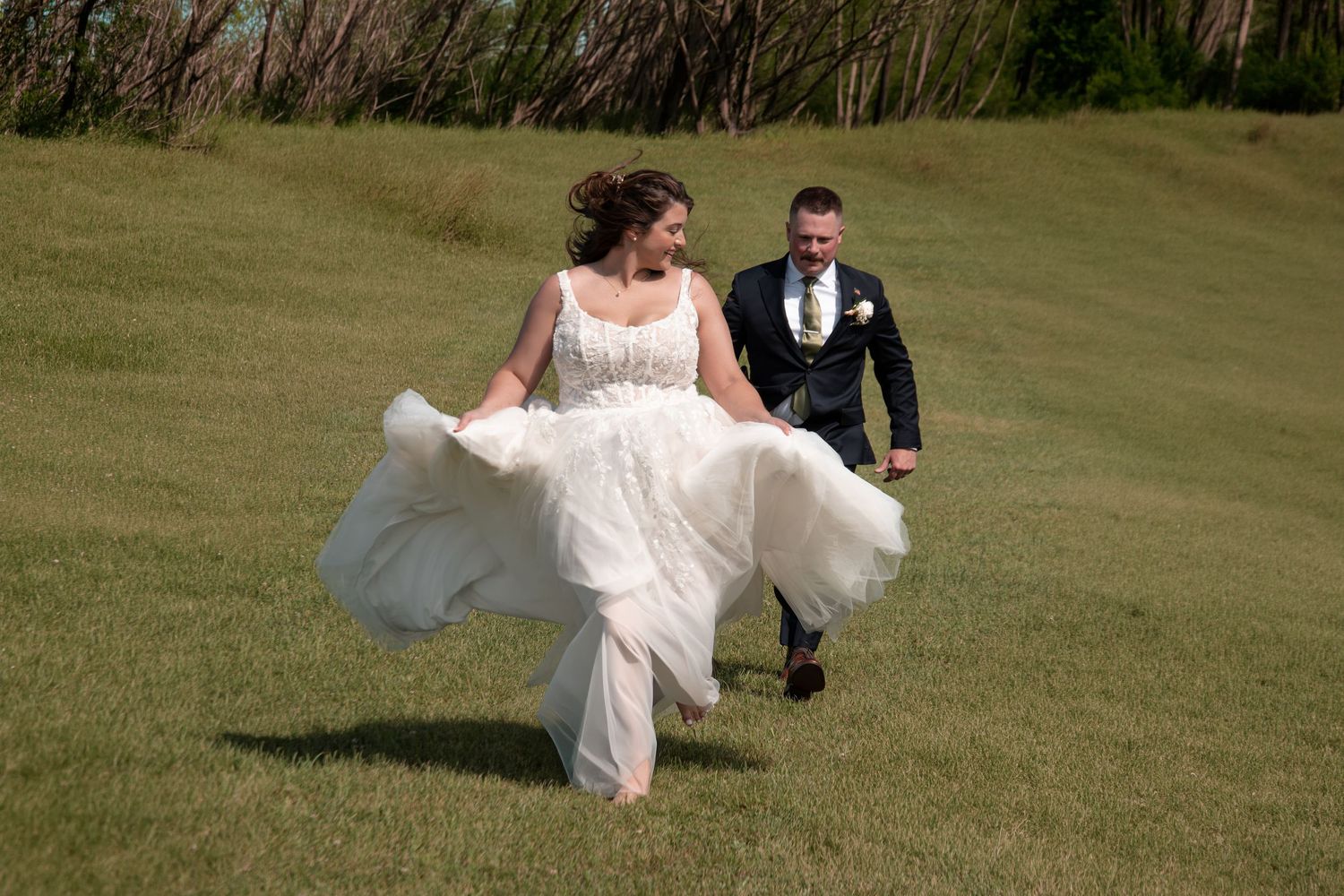 Bride in flowing white gown and groom in black suit running joyfully across green grass field on wedding day.