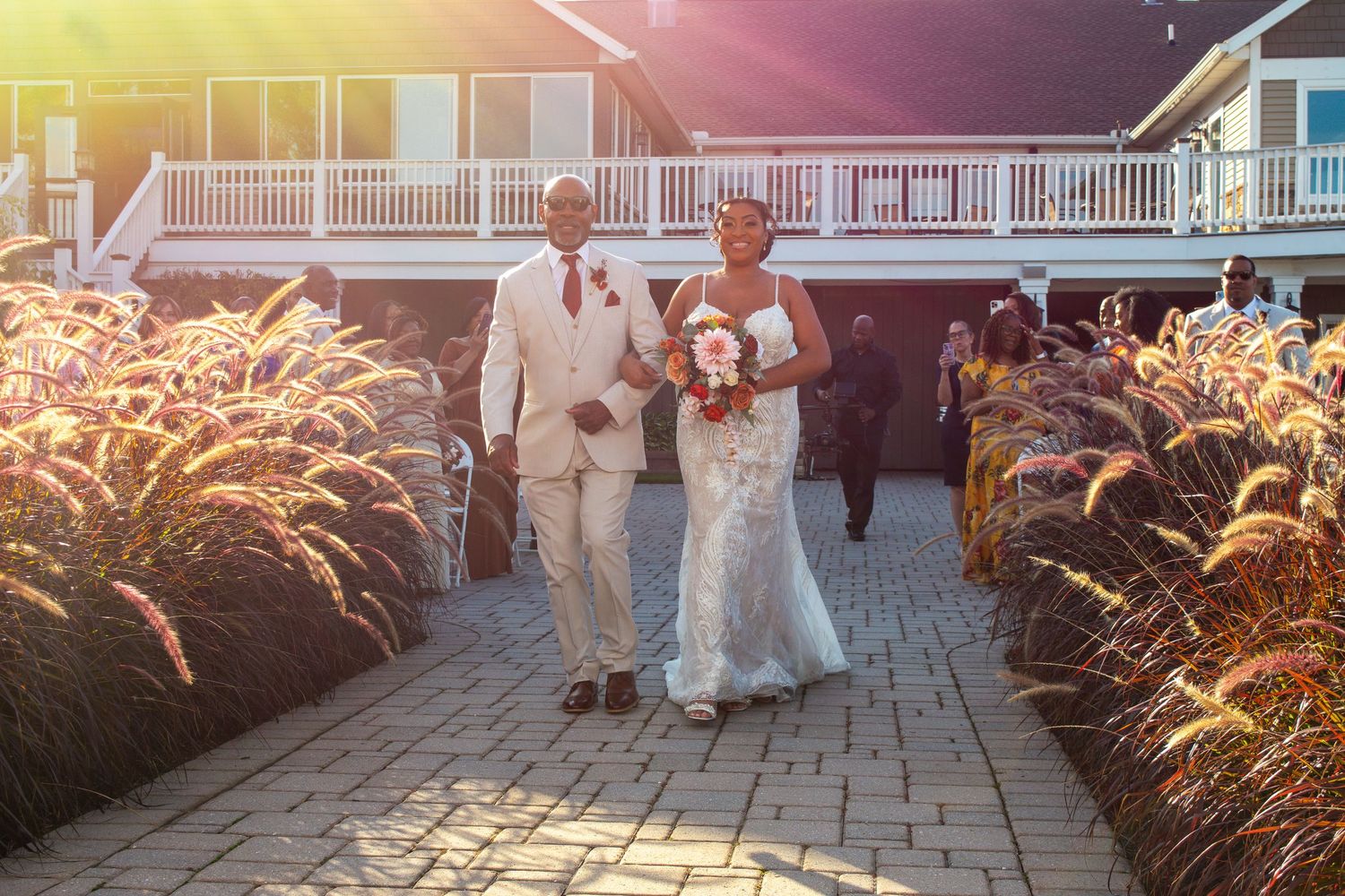 Bride and groom walking down outdoor aisle lined with ornamental grasses at golden hour wedding venue.
