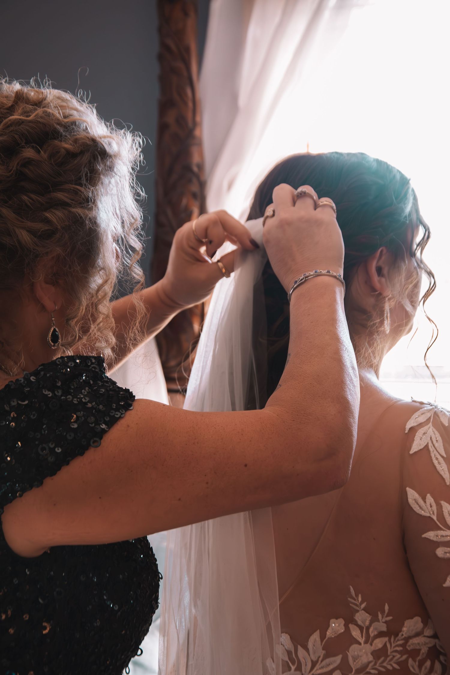 Mother helping bride secure wedding veil in hair during bridal preparations before ceremony.